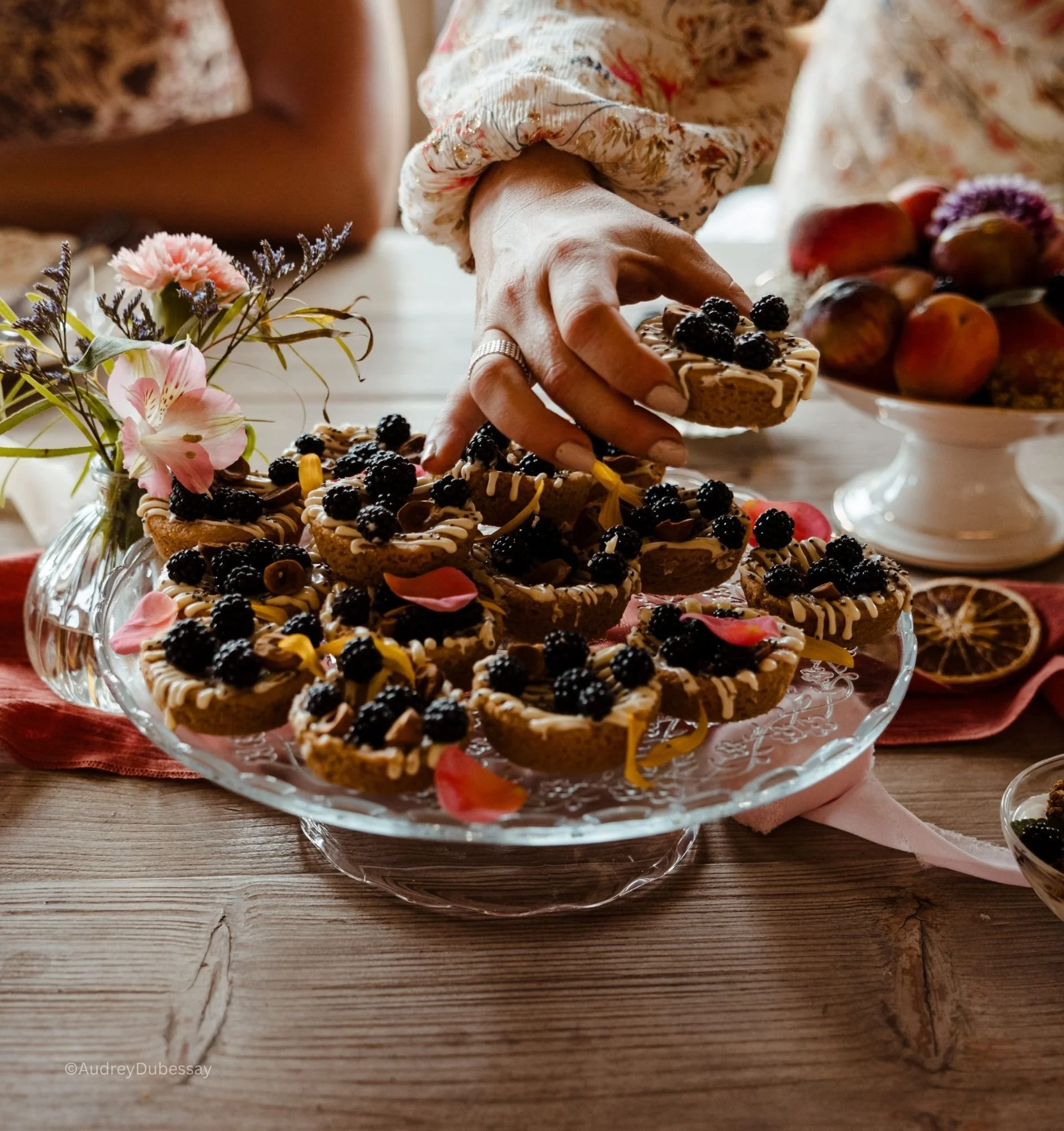 Une main féminine dépose des petits desserts au chocolat garnis de mûres noires, sur un plat en verre, avec des fleurs et une coupe de fruits en arrière-plan.