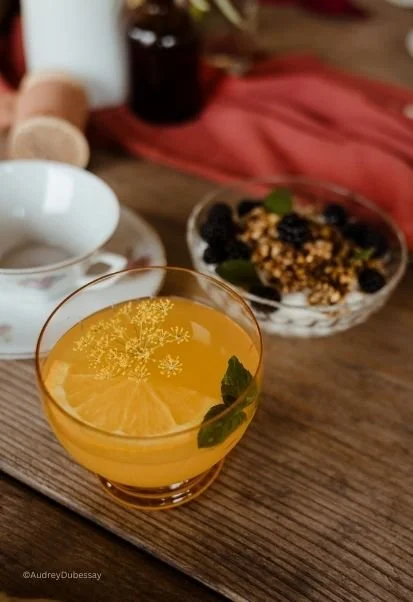 Verre d'une boisson orange avec une tranche de citron et une feuille de menthe, sur une table en bois avec un bol de fruits et une tasse blanche en arrière-plan.