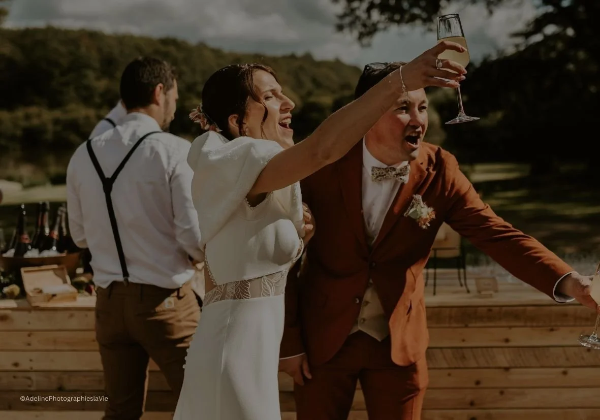 Un couple de mariés lors d'une célébration en plein air, le marié en costume marron et la mariée en robe blanche, trinant avec des verres de champagne, devant un décor naturel avec un fond de végétation.