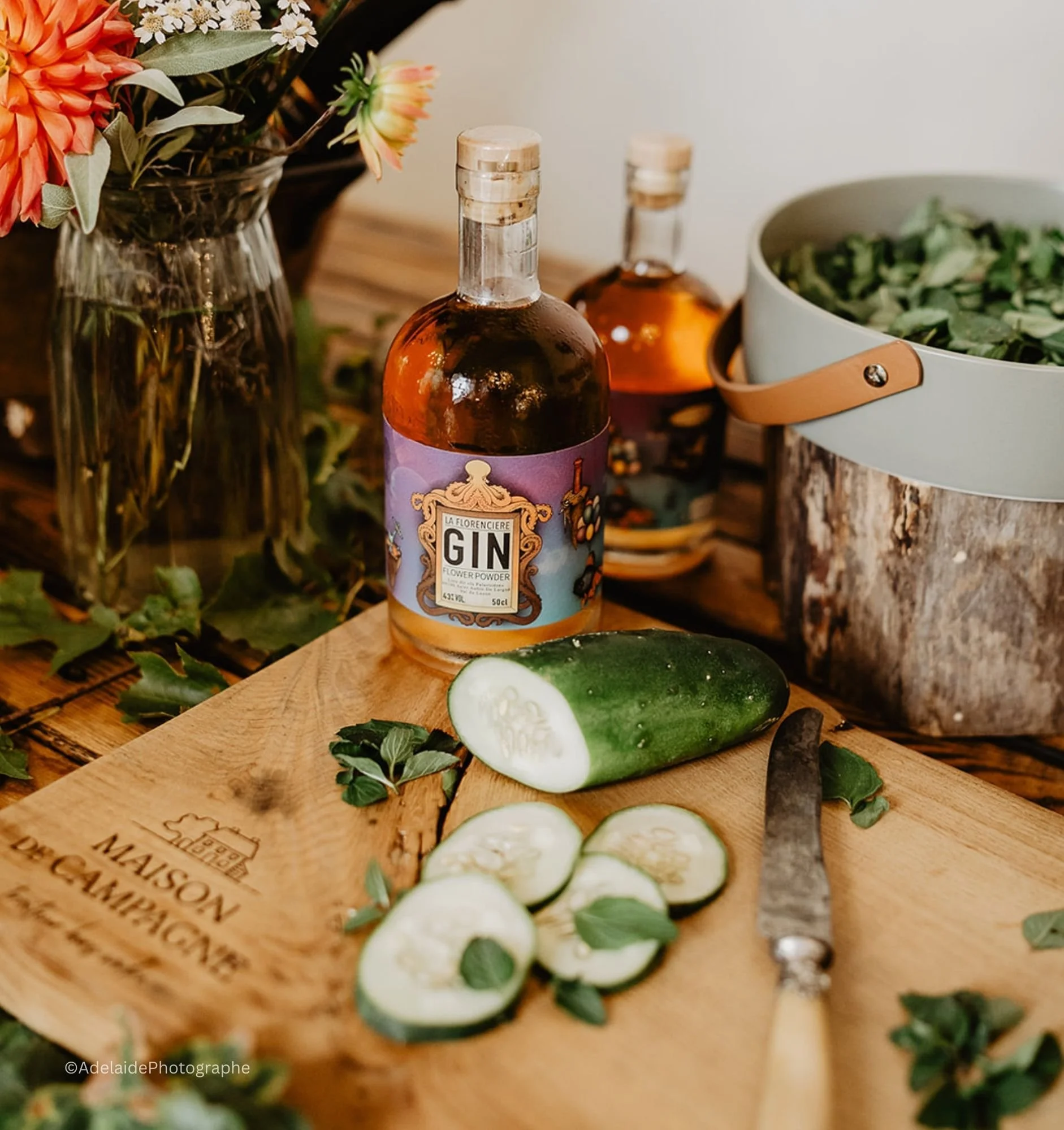Planche en bois avec concombre tranché, un couteau, et des feuilles d'herbes, entourée de bouteilles de gin, un vase de fleurs, et une boîte contenant des feuilles vertes.