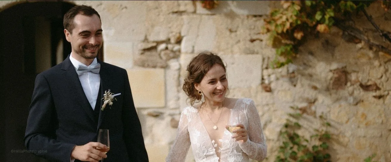 Un marié et une mariée portant une robe blanche délicate, tenant des flûtes de champagne lors d'une célébration en plein air, avec un mur en pierre en arrière-plan.