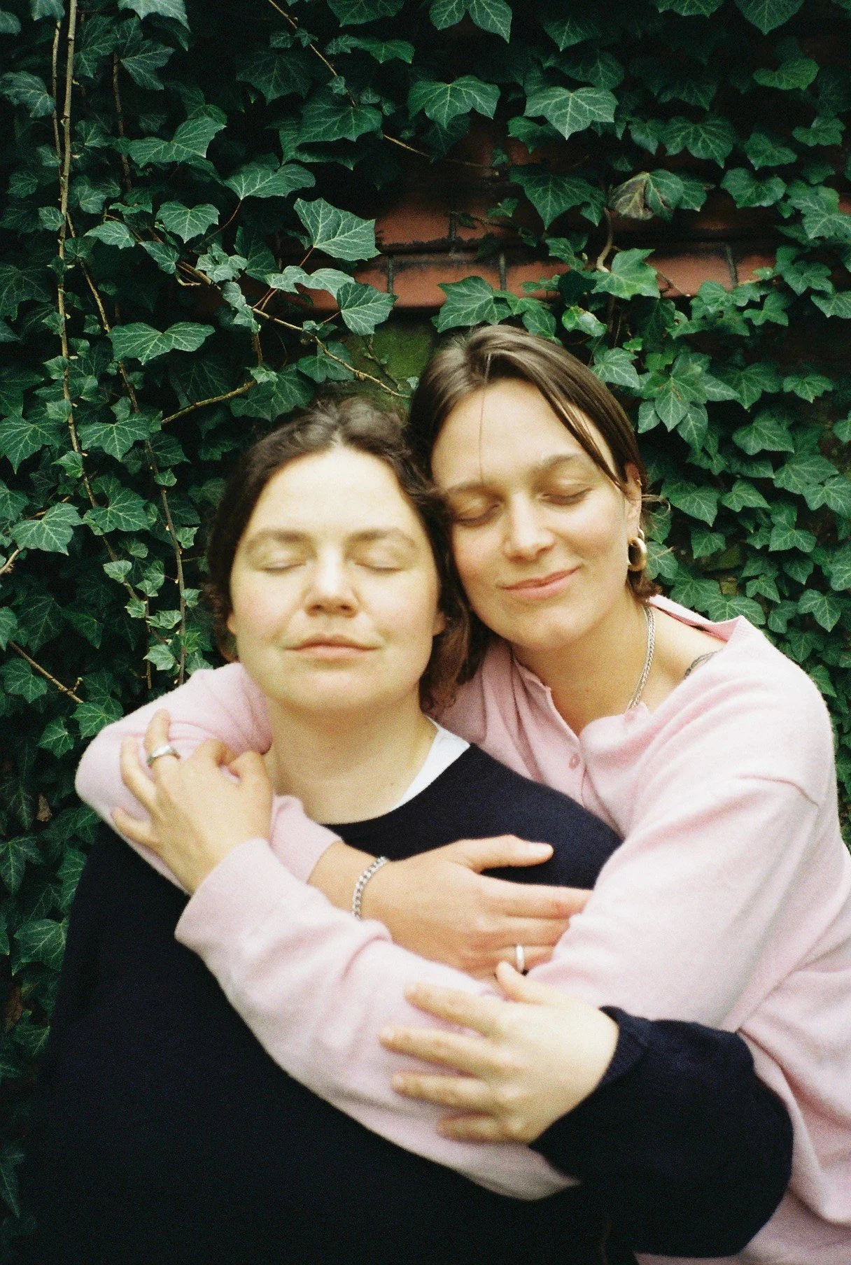 Zwei Frauen umarmen sich liebevoll vor einem grünen, bepflanzten Hintergrund, beide haben die Augen geschlossen und lächeln.