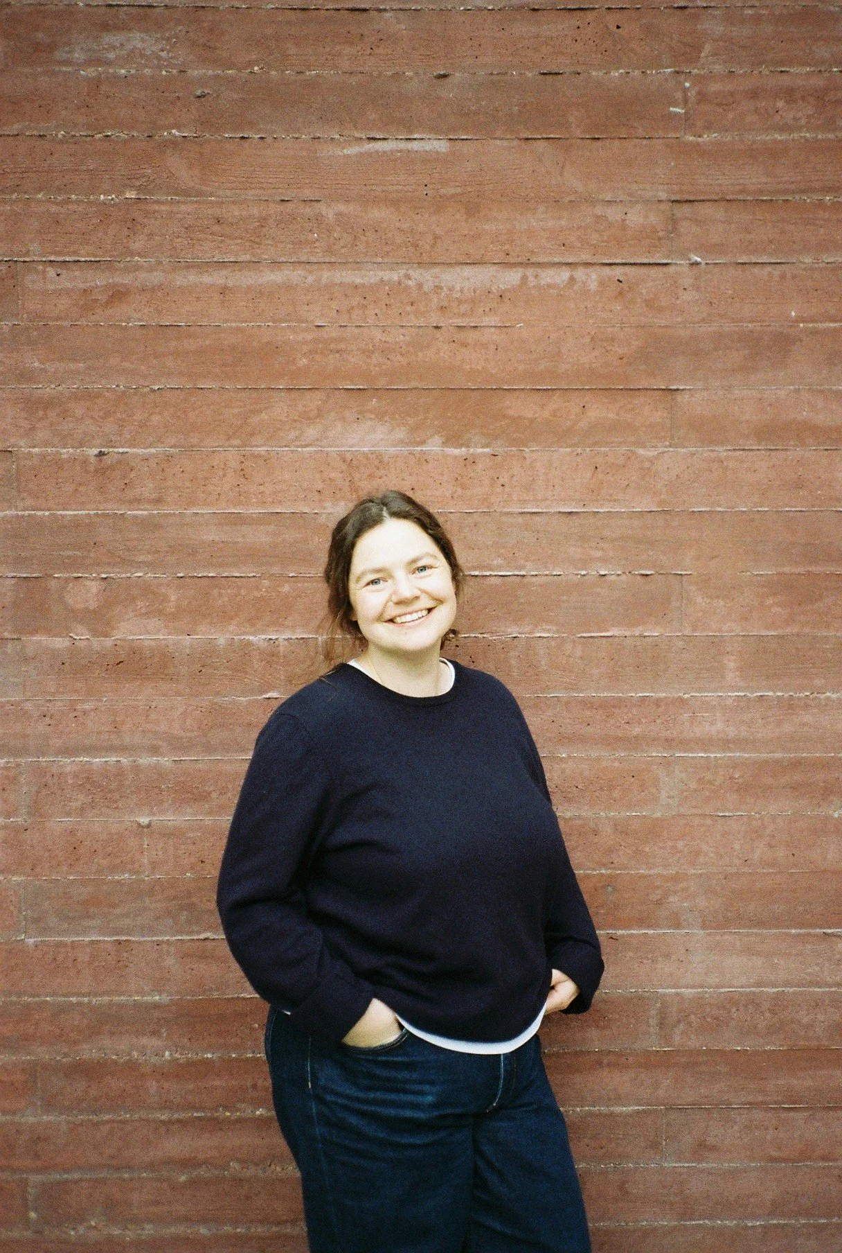 Eine lächelnde Frau mit braunen, glatten Haaren, die eine schwarze Pullover und Jeans trägt, posiert vor einer roten Backsteinmauer.