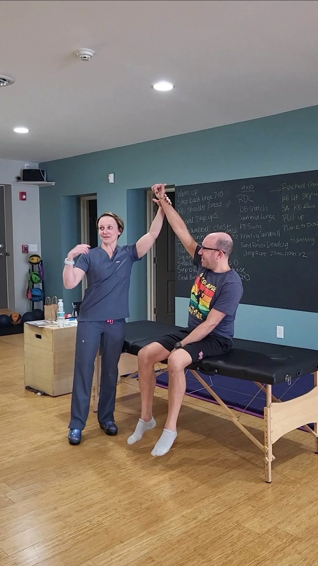 Angela of North Country Acupuncture demonstrates on a man sitting on a medical examination table and stretching his arm.