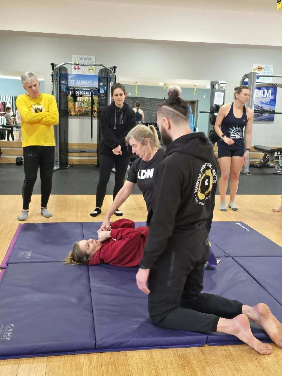 A woman lies on a padded mat practicing self-defense techniques with a partner kneeling beside her. Master Kyle Phillips demonstrating martial arts techniques to a group of women and children at Keep Fit in Guilderland, NY.