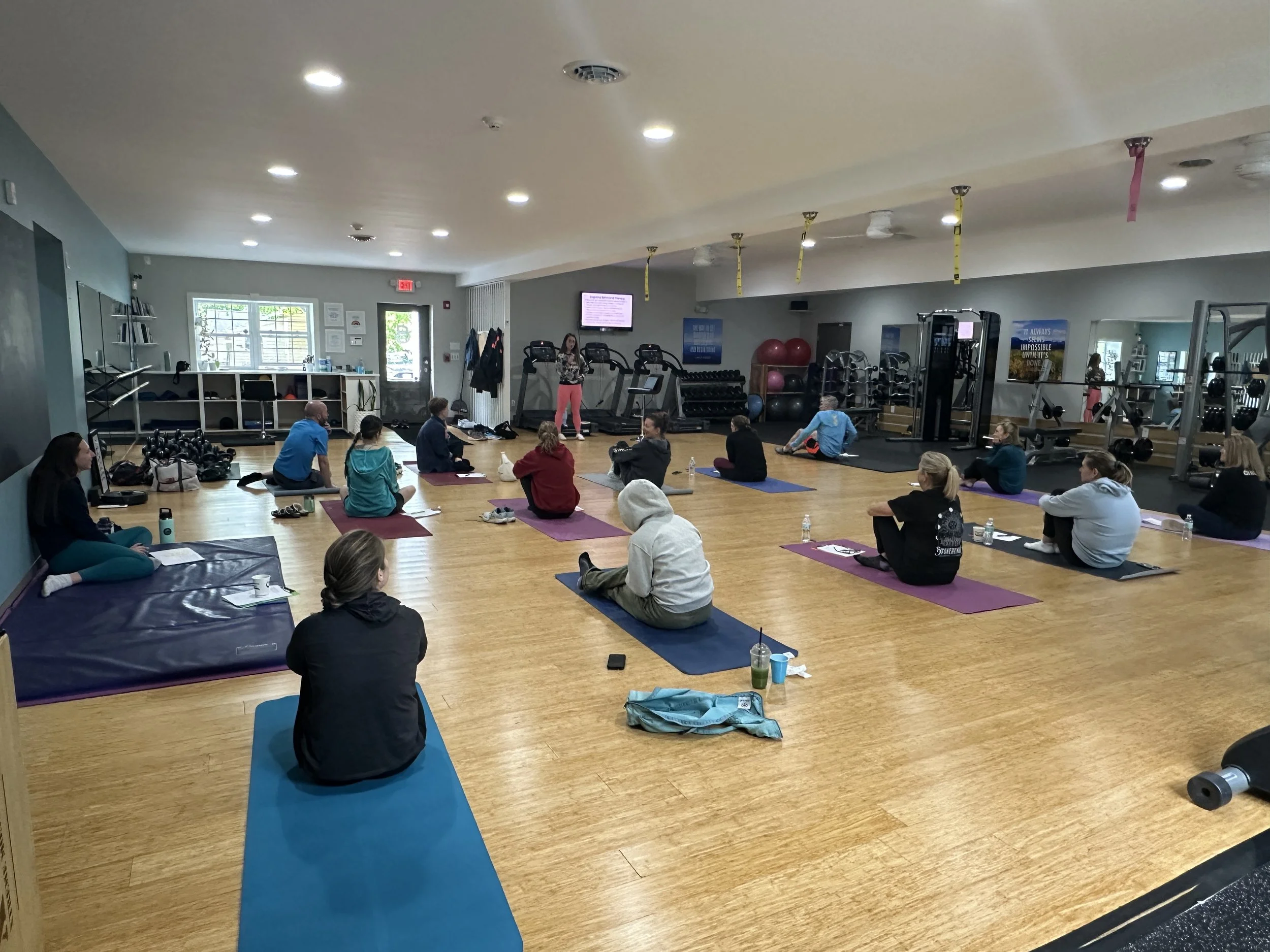 Indoor yoga or fitness class with participants seated on mats, instructor at the front near treadmill machines, mirrors on the wall, and exercise equipment in the background.