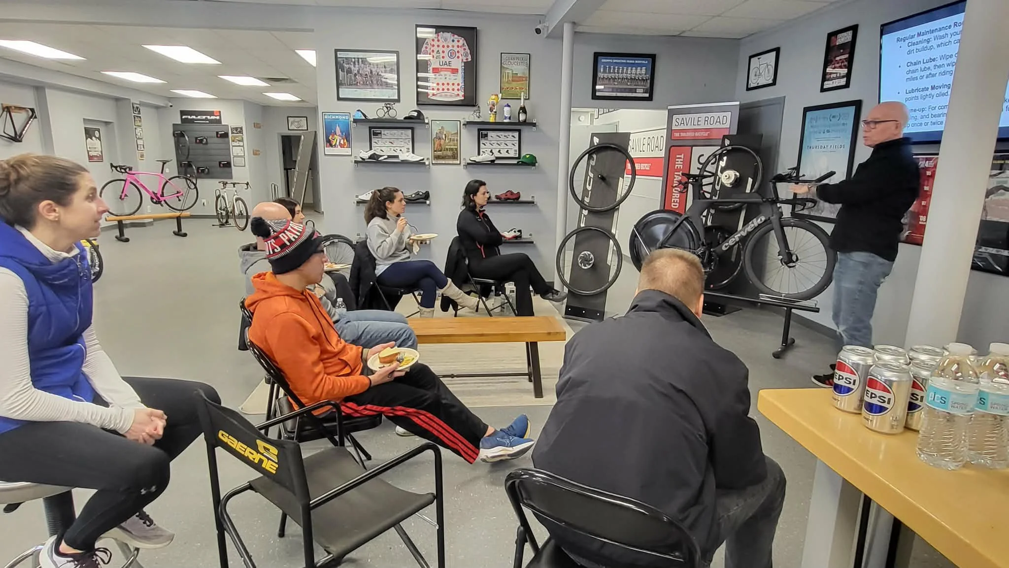 A group of people attending a bicycle maintenance seminar in a bike shop, with a screen displaying maintenance information. The audience is seated and listening attentively.