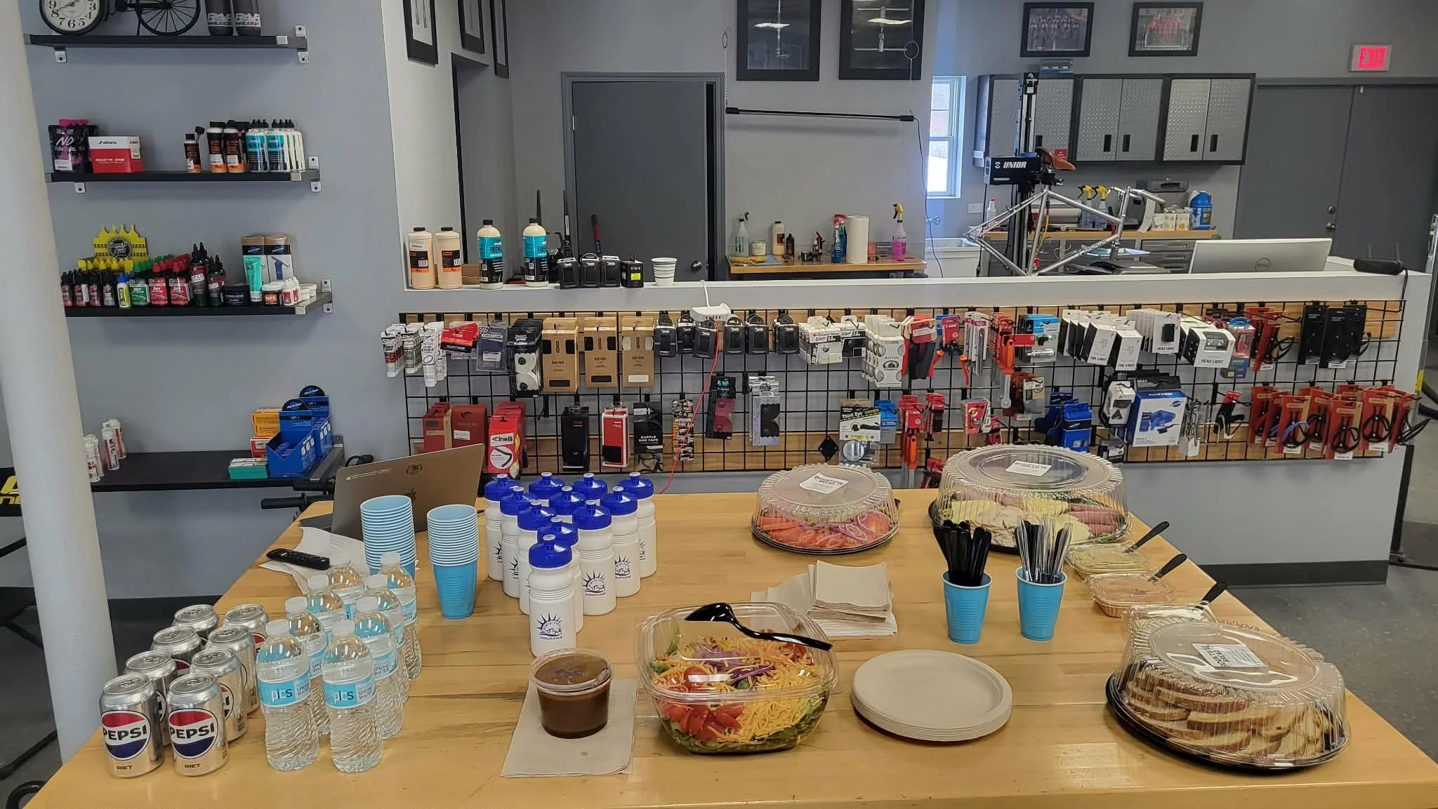 A table with bottled water, cans of Pepsi, salad, and desserts in a room with shelves and a large sign in the background
