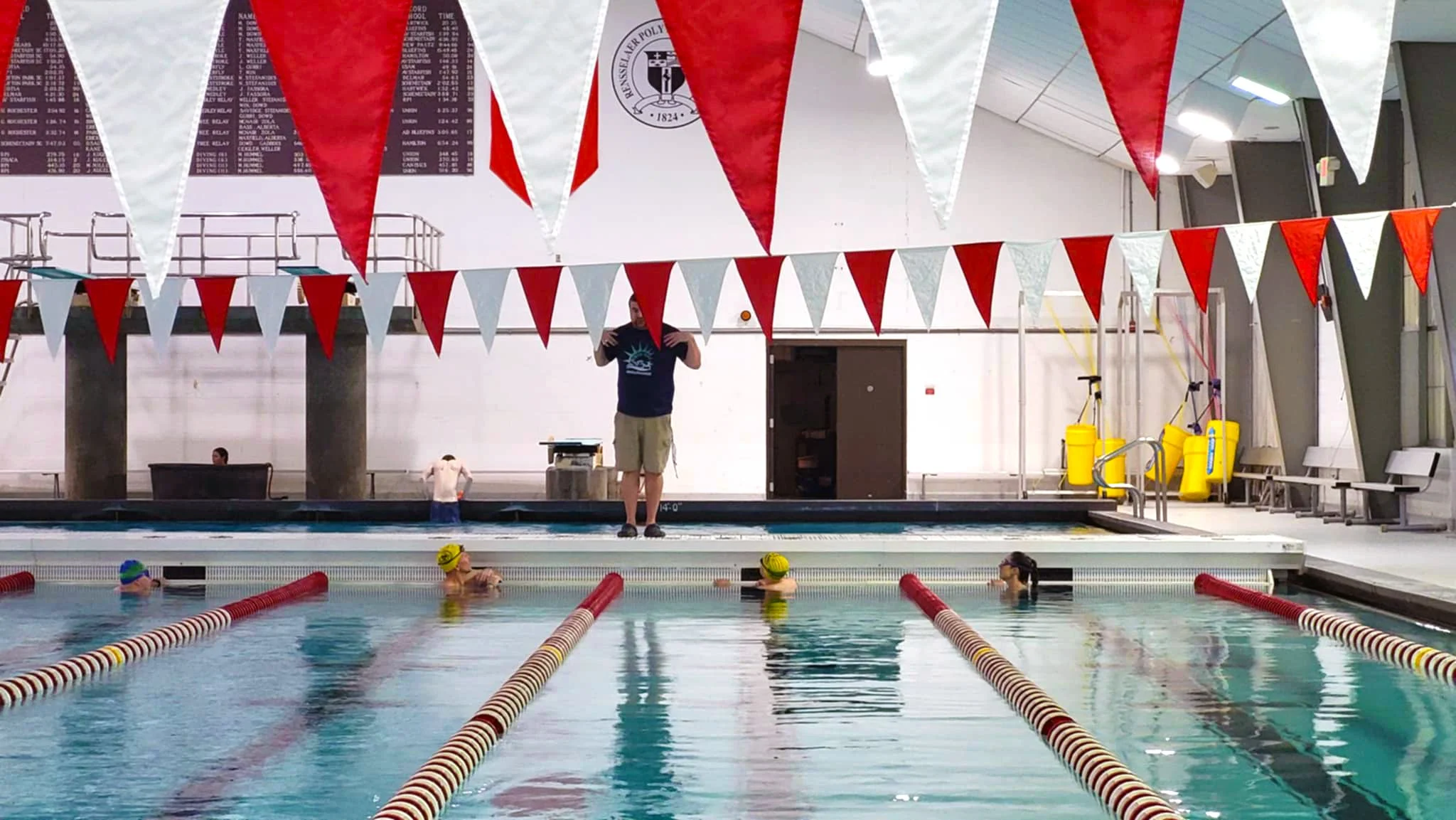 Indoor swimming pool with triathletes wearing swim caps, swimming in lanes, and an adult coach or instructor standing on the pool deck.