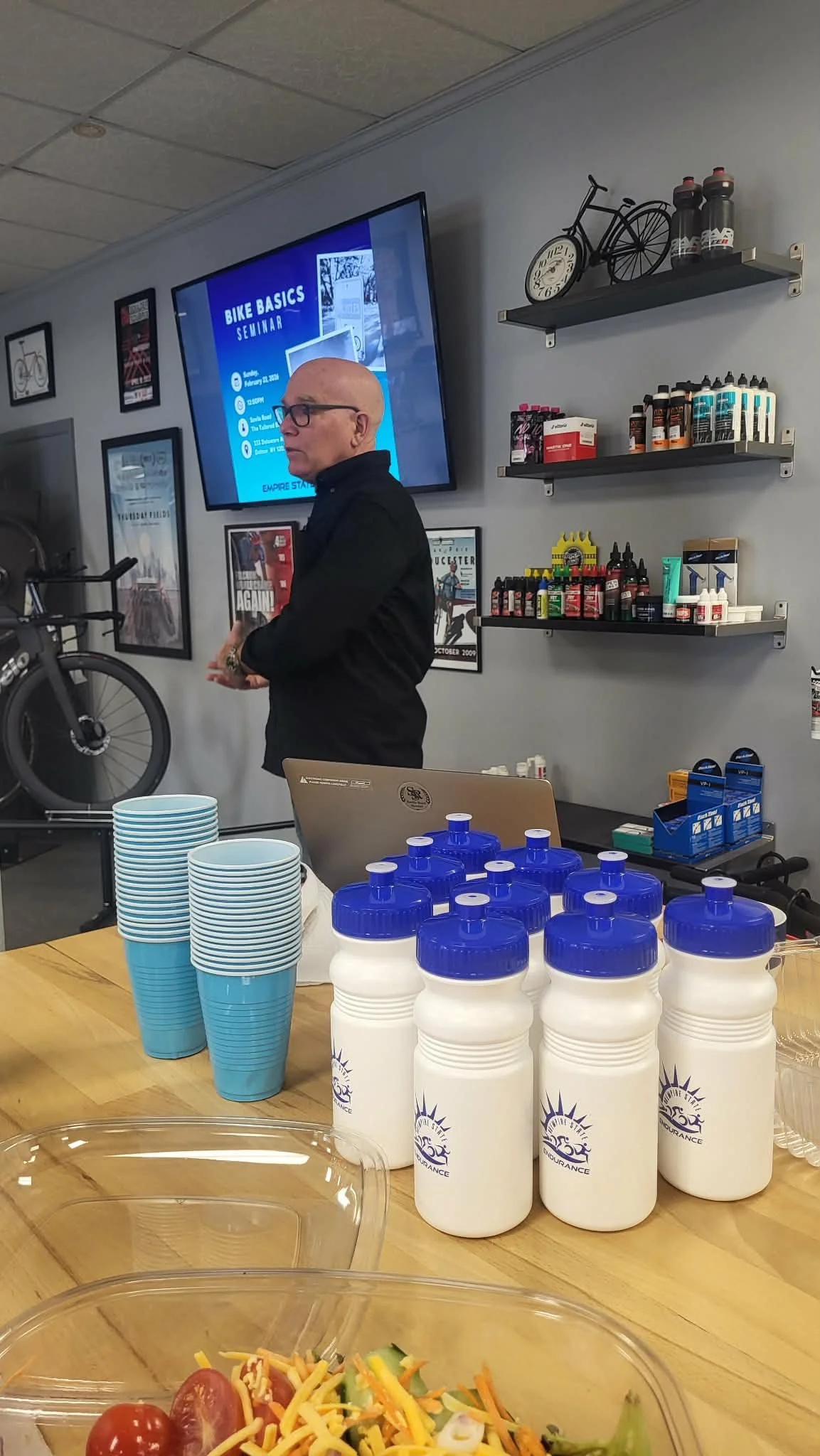 Cafe table with white water bottles with blue caps, blue cups, and salad containers, with a man standing behind a laptop and a presentation screen about bike basics seminar, walls decorated with framed posters, bicycle and supplies on shelves.