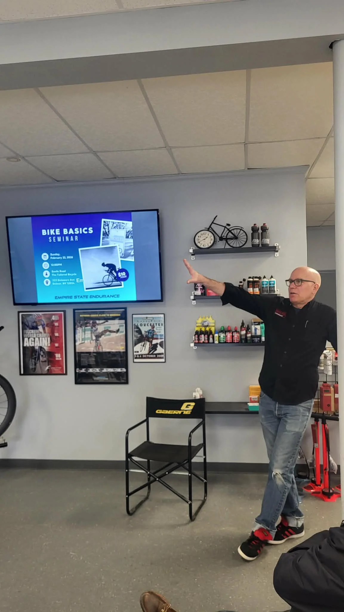 Steve of Saville Road, the tailored bicycle, is giving a presentation in a room with bicycle-themed decor. A television screen displays an announcement for a bike basics seminar.