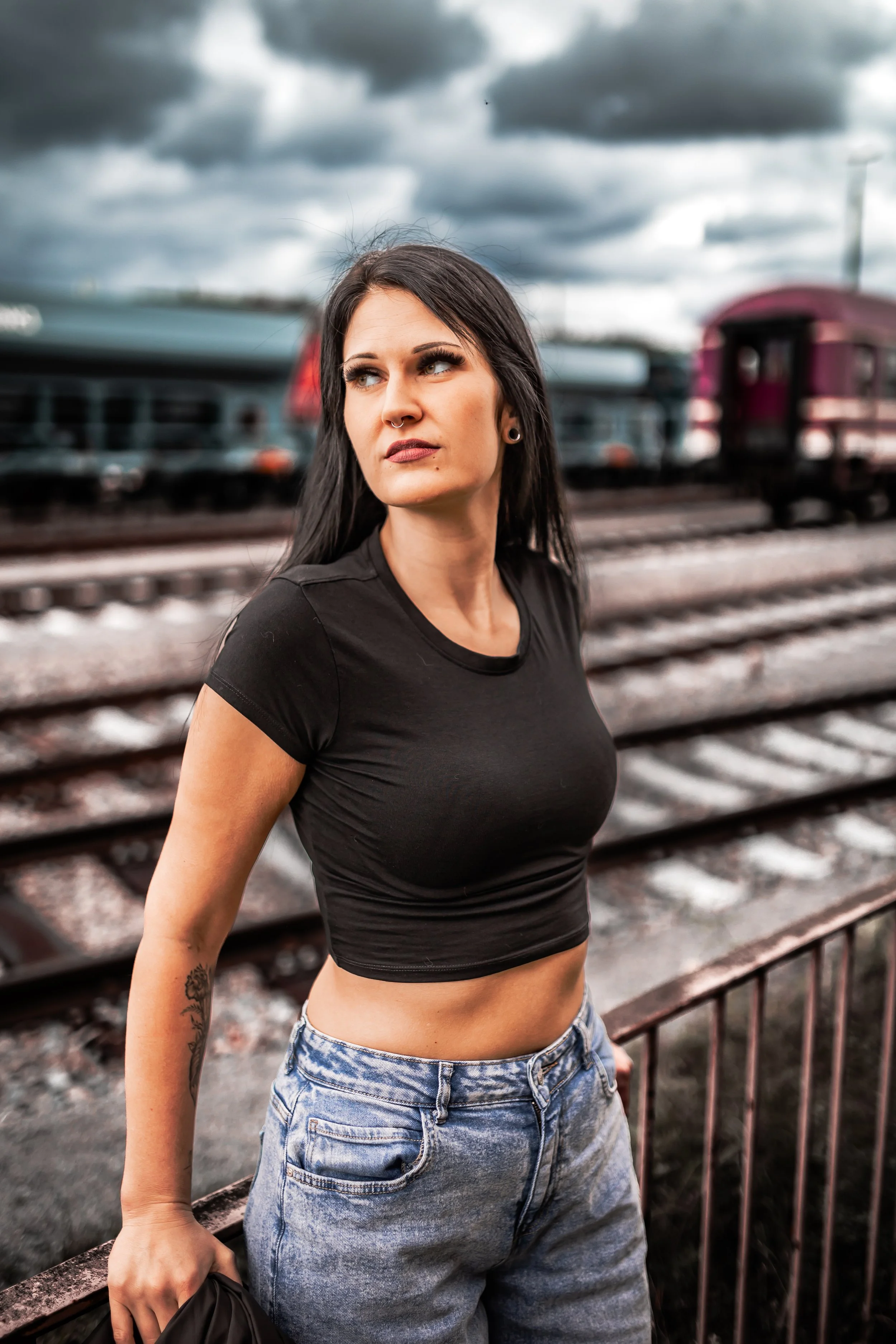 Junge Frau steht am Bahnsteig vor Zügen bei schlechten Wetter, trägt schwarzes Crop-Top und Jeans, hat dunkle Haare und sichtbare Tätowierungen am Arm.