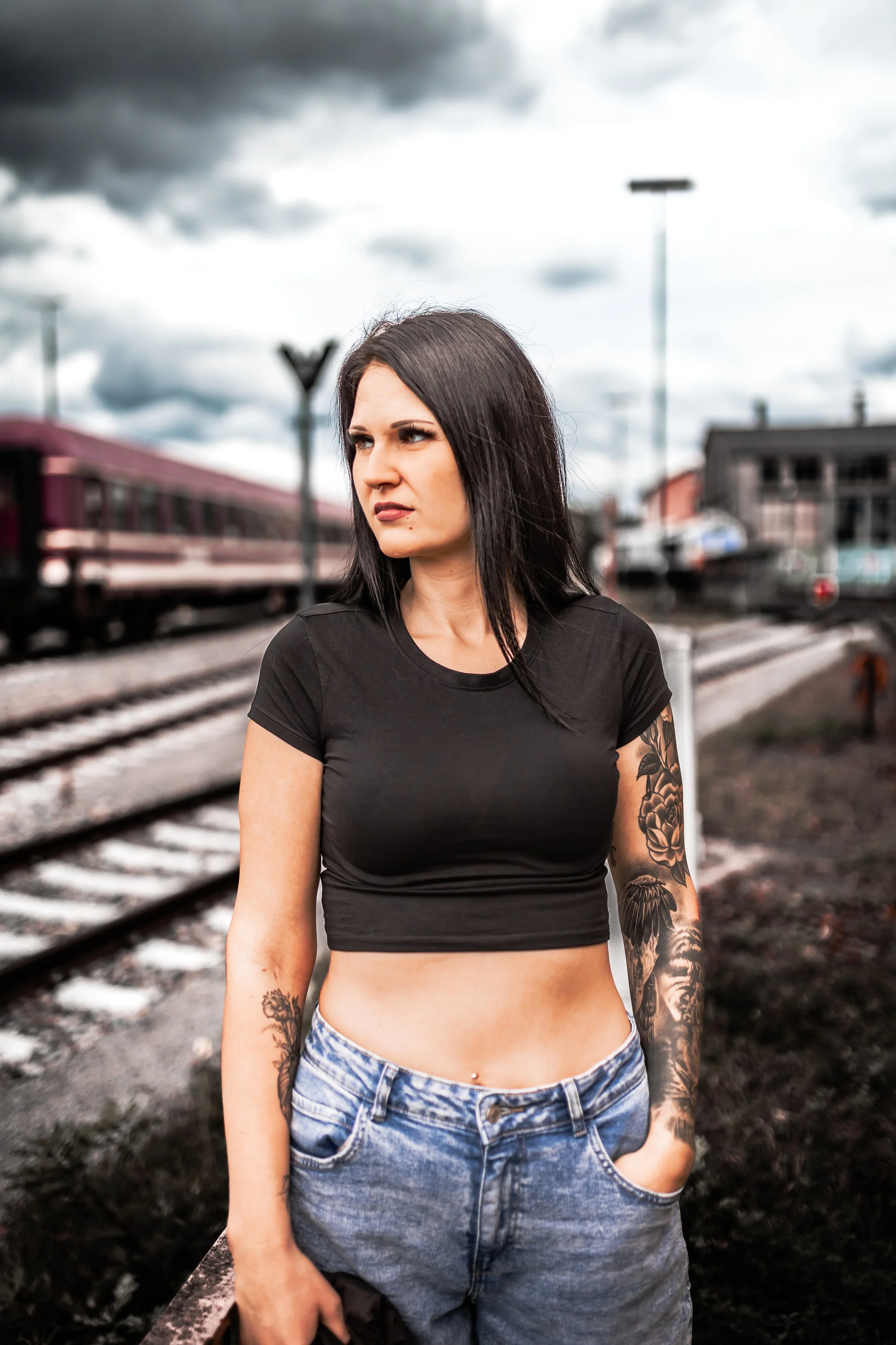 Junge Frau steht auf einem Bahnsteig bei bewölktem Himmel, trägt ein schwarzes Crop-Top und Jeans, hat Tattoos und schwarze Haare.