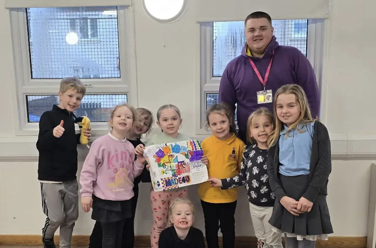 Group of children and an adult posing indoors with a colorful poster, one child holding a banana.