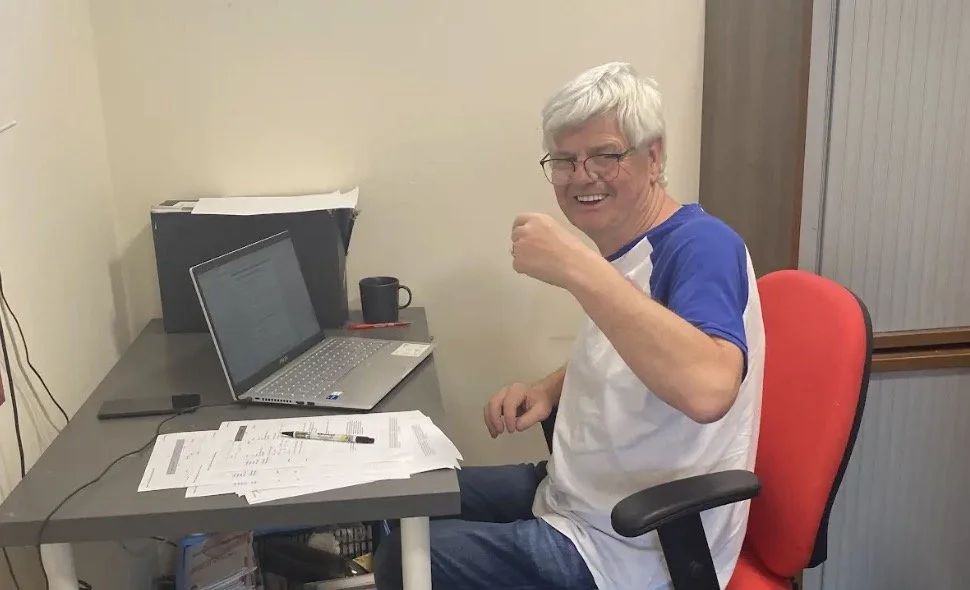 Person sitting at a desk with a laptop and papers, smiling and giving a thumbs up.