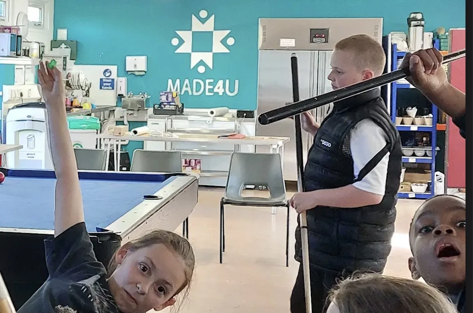 Children playing pool in a recreation room with a pool table, kitchen area, and various supplies in the background.