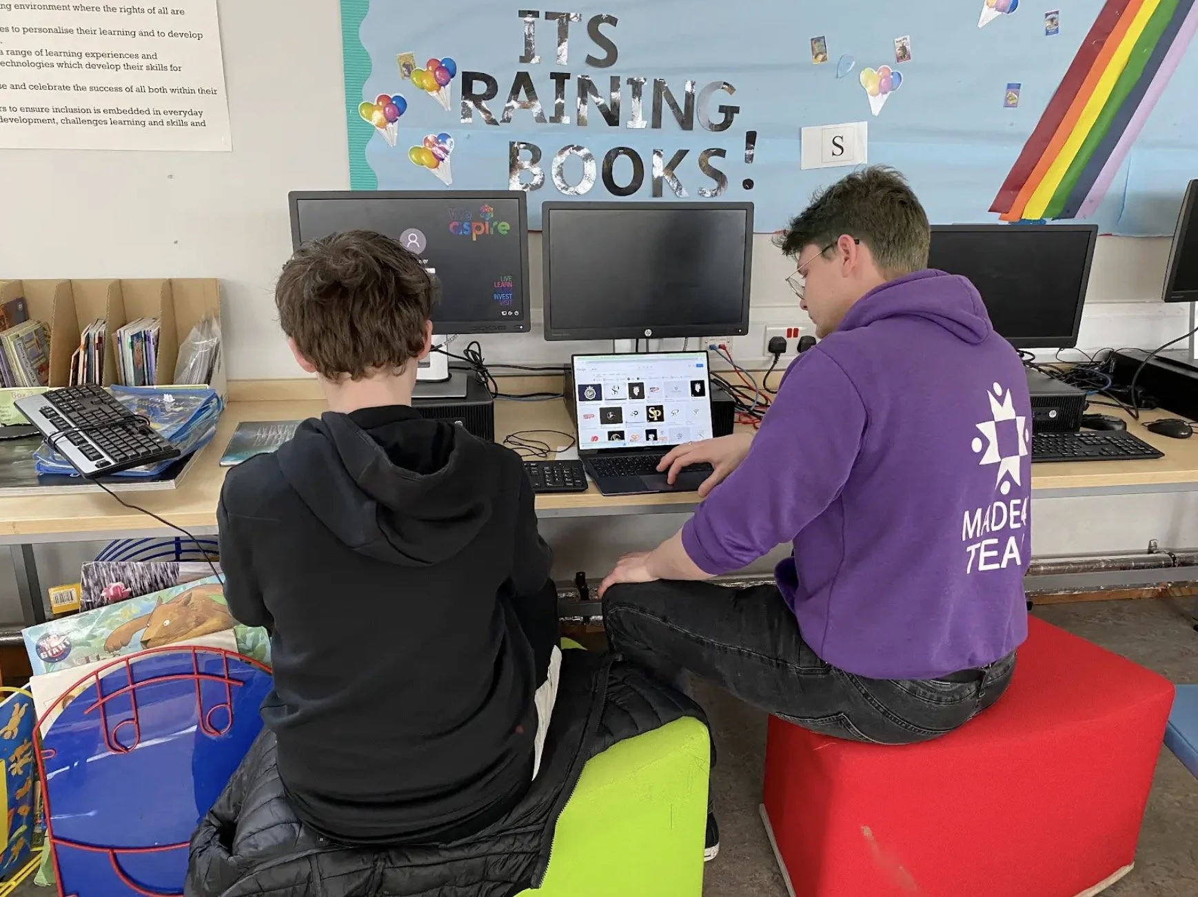 Two people sitting at a desk with computers, books, and a laptop, under a bulletin board that reads 'It's Raining Books!' with balloons and a rainbow decoration.