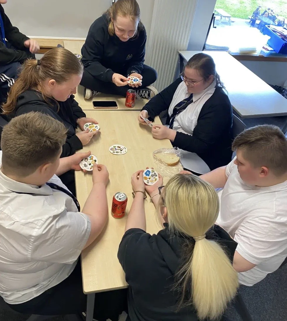 Group of people playing a card game at a table with soda cans and a sandwich.