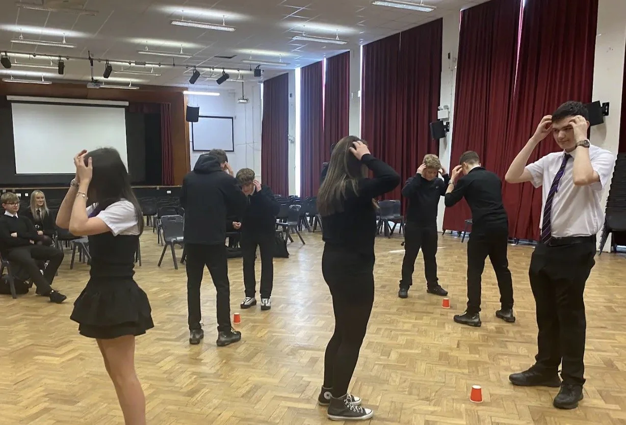 Students playing a game with plastic cups in a school hall, some standing with their hands on their heads and others sitting in chairs.
