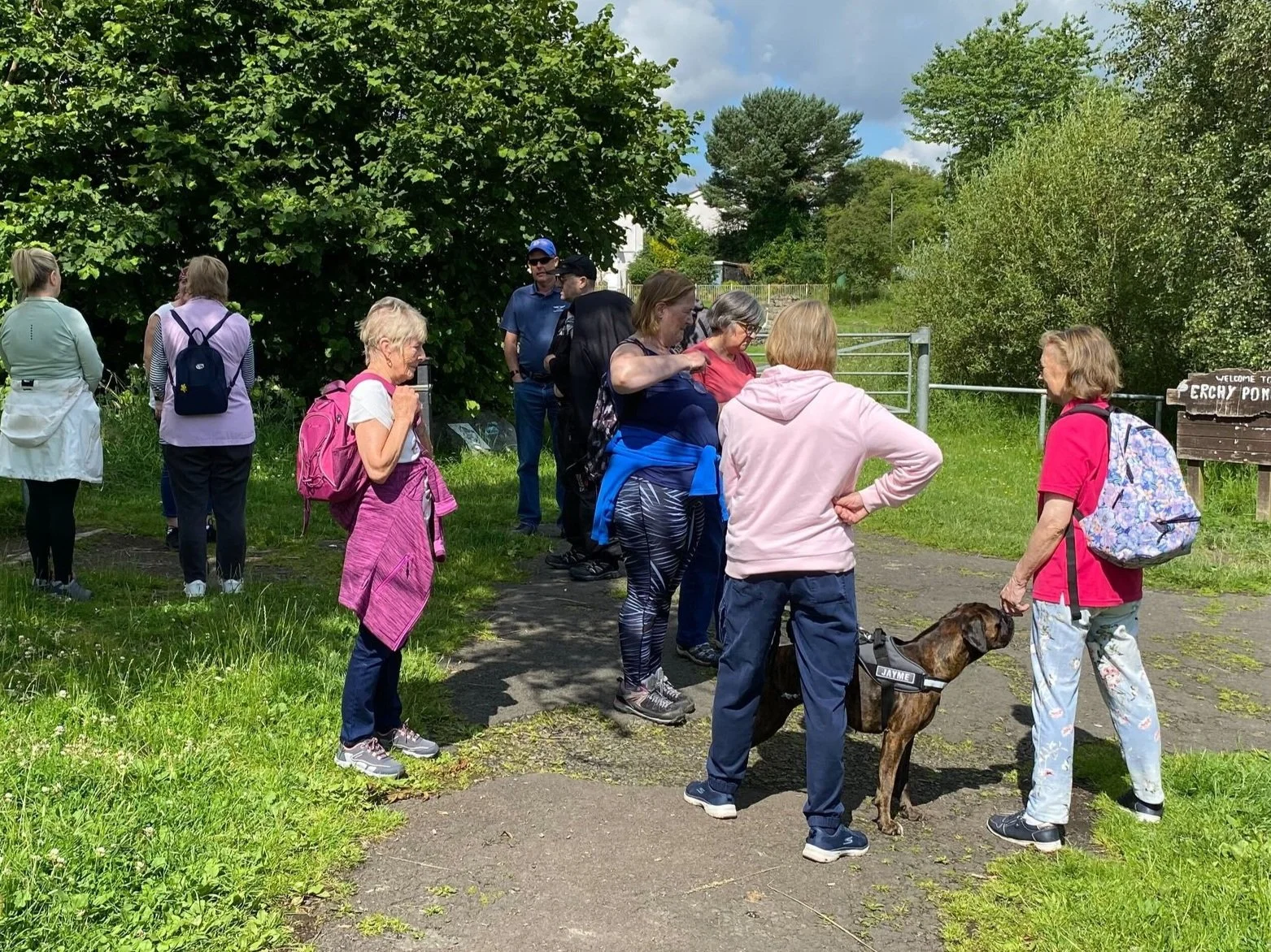 Group of people gathered in a park, some wearing backpacks, with a dog, greenery, and a sign reading "Perchy Pond" in the background.