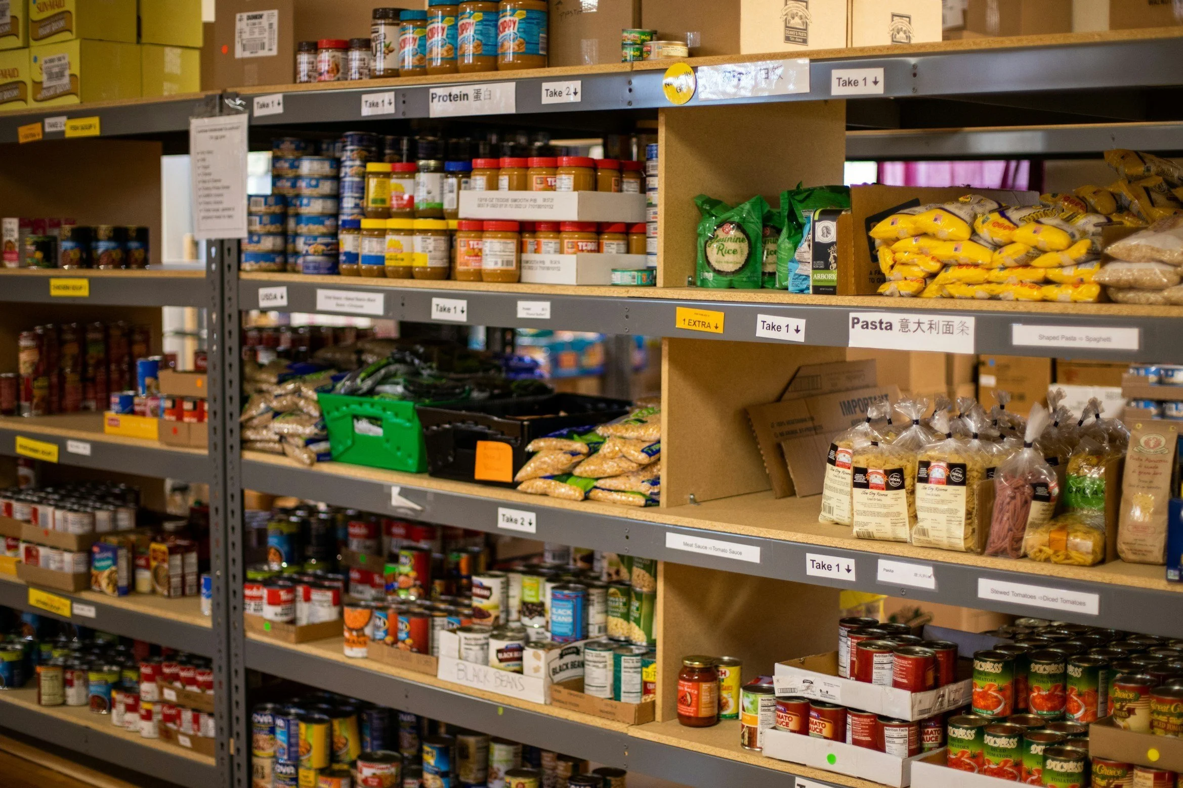 Shelves stocked with various canned and packaged food items, including pasta, rice, canned beans, peanut butter, and other non-perishable goods in a food pantry.