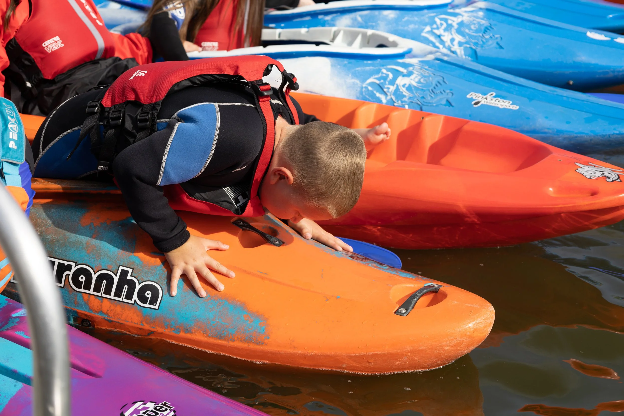 Child wearing a red life vest leaning over an orange kayak on water, surrounded by other colorful kayaks.