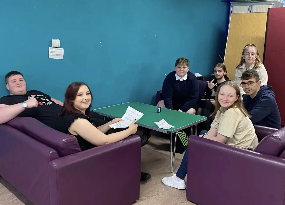 Group of people sitting around a table in a room with purple couches and a blue wall, one holding a paper.