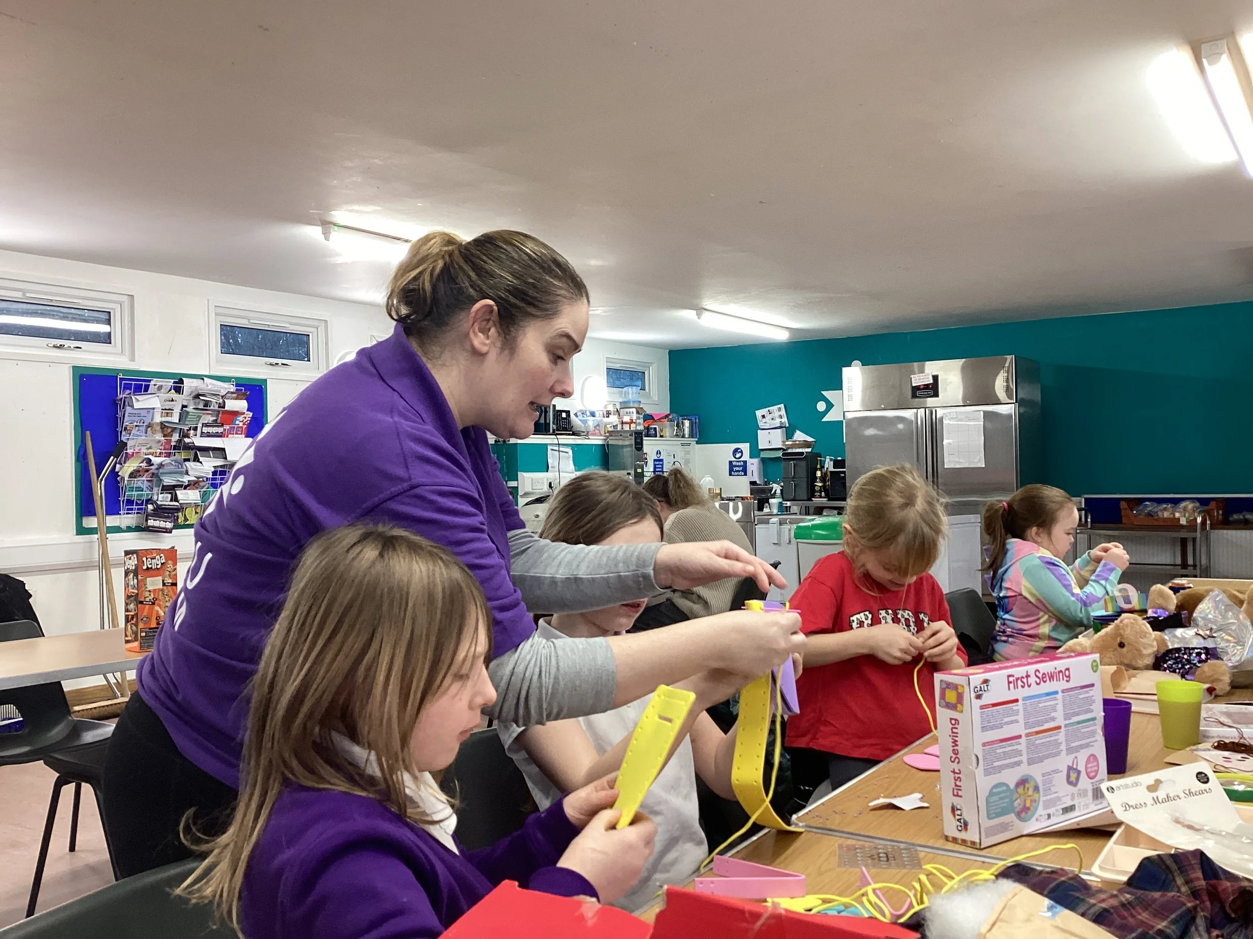 An instructor teaching children how to sew in a classroom with various sewing supplies and teddy bears on the table.