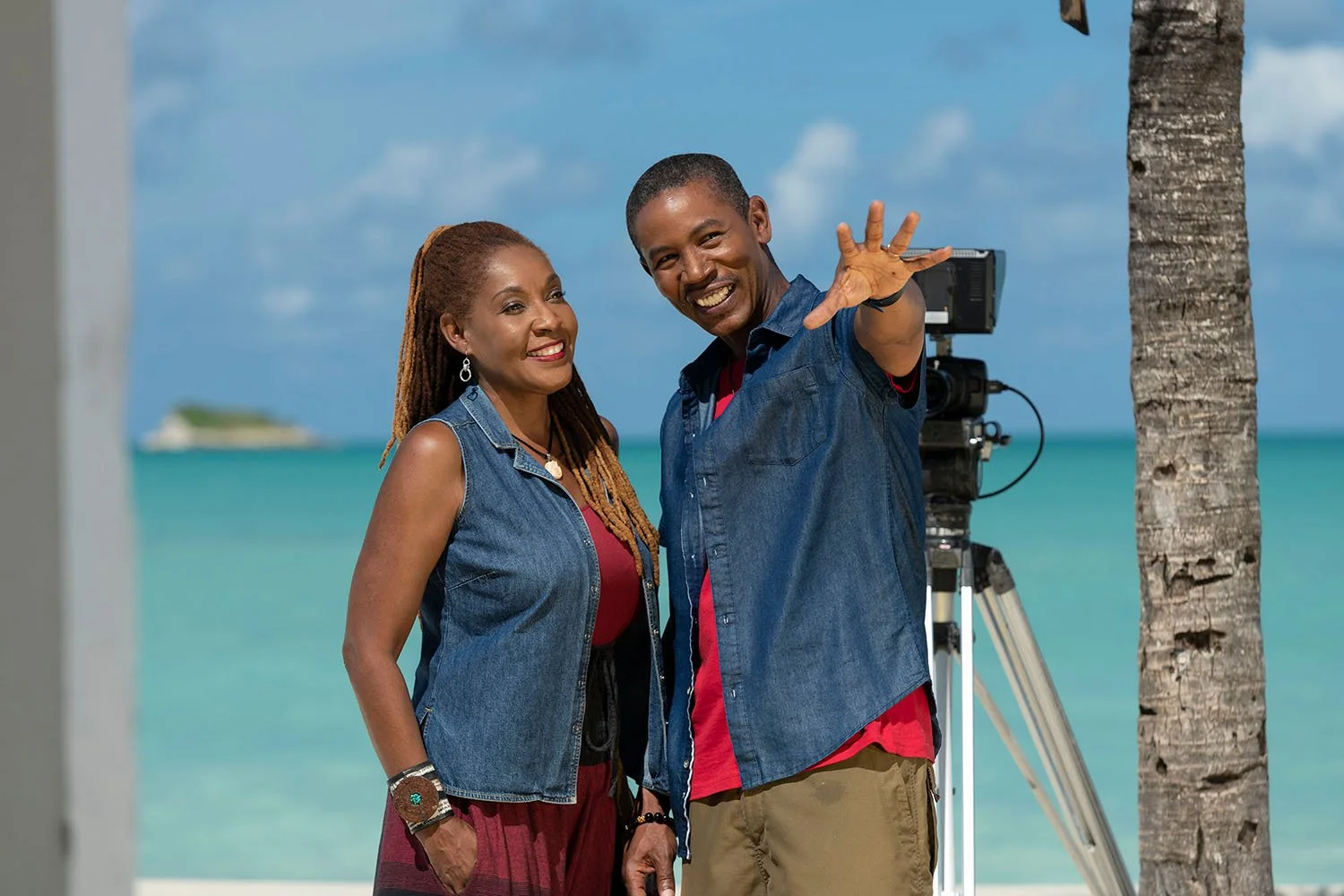 Two smiling people, a woman and a man, standing on a beach with turquoise water, palms, and a photographer's camera on a tripod behind them. The man is gesturing with his hand, and both are wearing denim vests.
