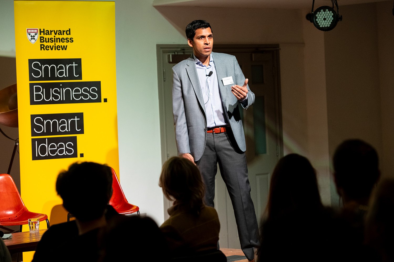 A man giving a presentation on stage at a Harvard Business Review event, with a yellow banner that reads 'Smart Business. Smart Ideas.' and a small audience watching.