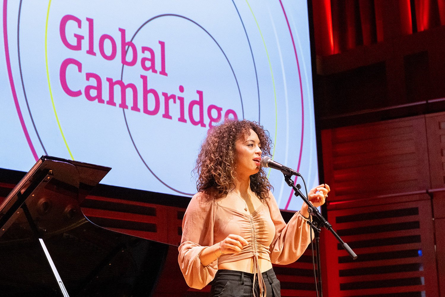 A woman with curly hair speaking into a microphone on stage at an event, with a large screen behind her displaying the logo 'Global Cambridge'.