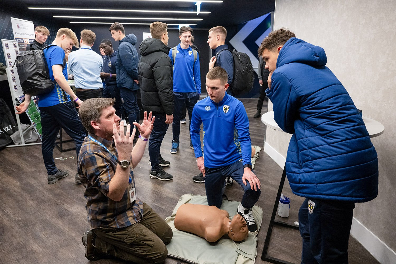 Soccer players in blue training gear practicing CPR and first aid techniques on a mannequin during a training session in an indoor facility.