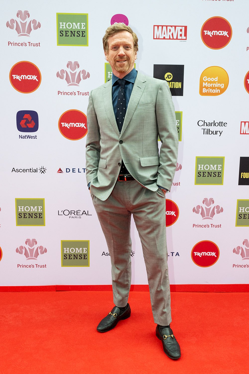 A man in a light gray suit standing on a red carpet at an event, with logos of various sponsors and organizations behind him.