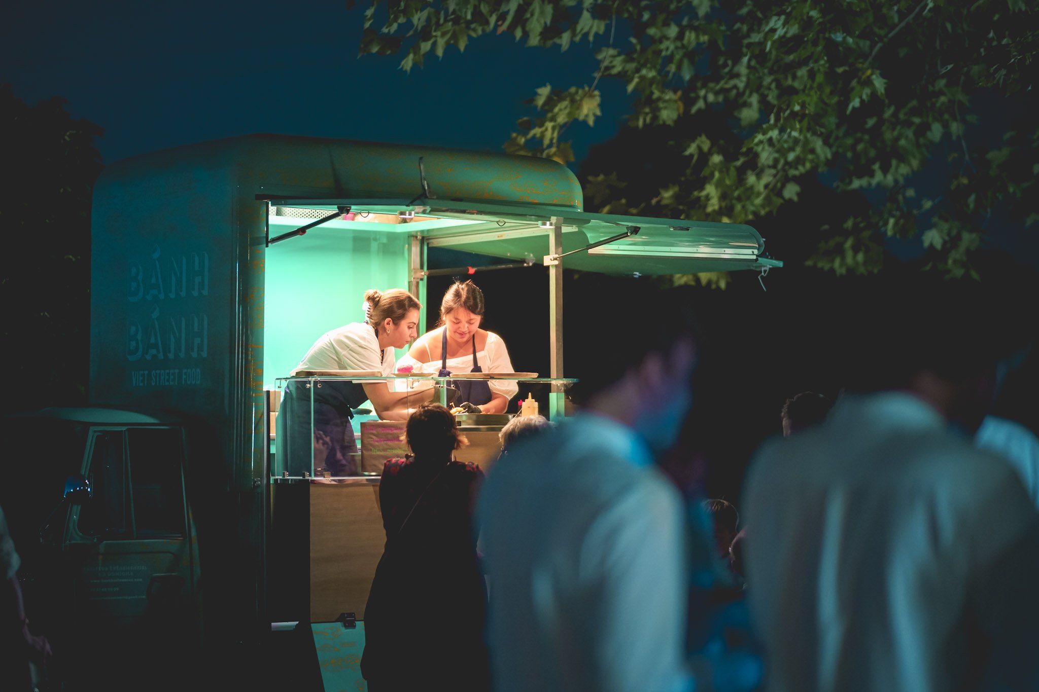 Un food truck de cuisine vietnamienne avec deux personnes préparant de la nourriture, entourées de clients en soirée.