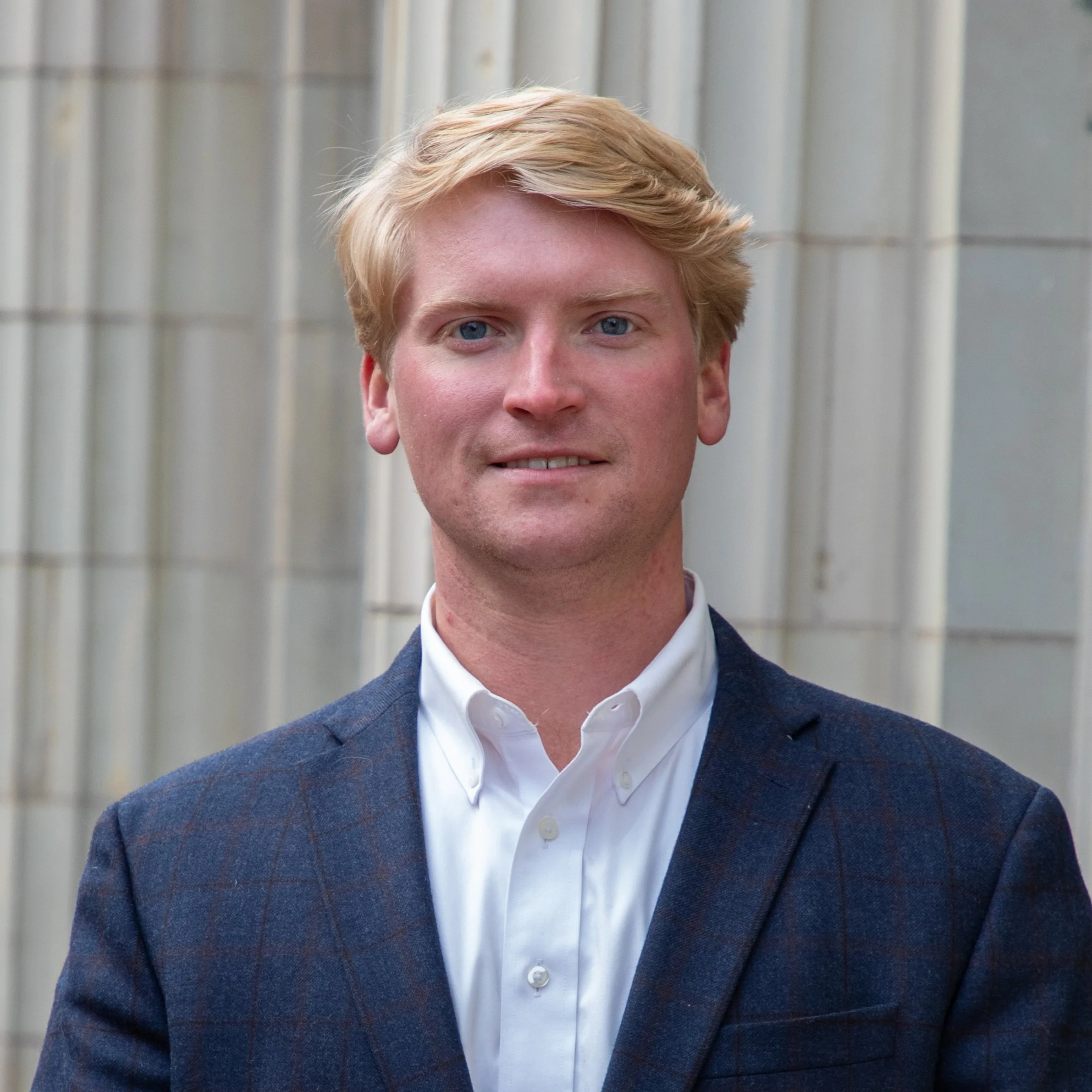 Portrait of a young man with blonde hair wearing a white shirt and a dark blazer, standing outdoors in front of a building with metal siding.