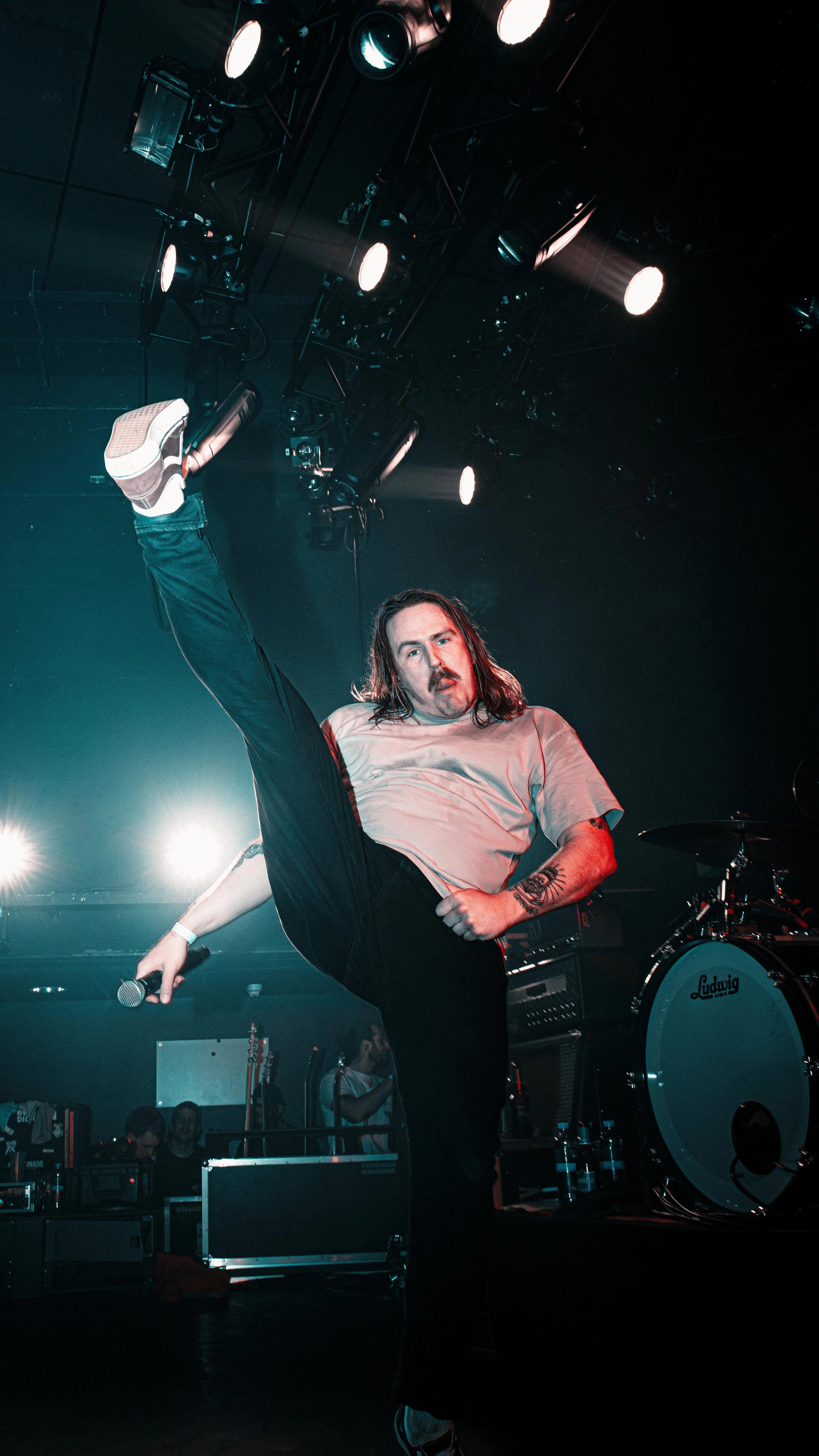 A male performer with long hair, mustache, and tattoos, wearing a gray T-shirt and black pants, stands on stage with one leg raised high in a kick, holding a microphone in his left hand. The stage has several bright lights overhead, and a Ludwig drum
