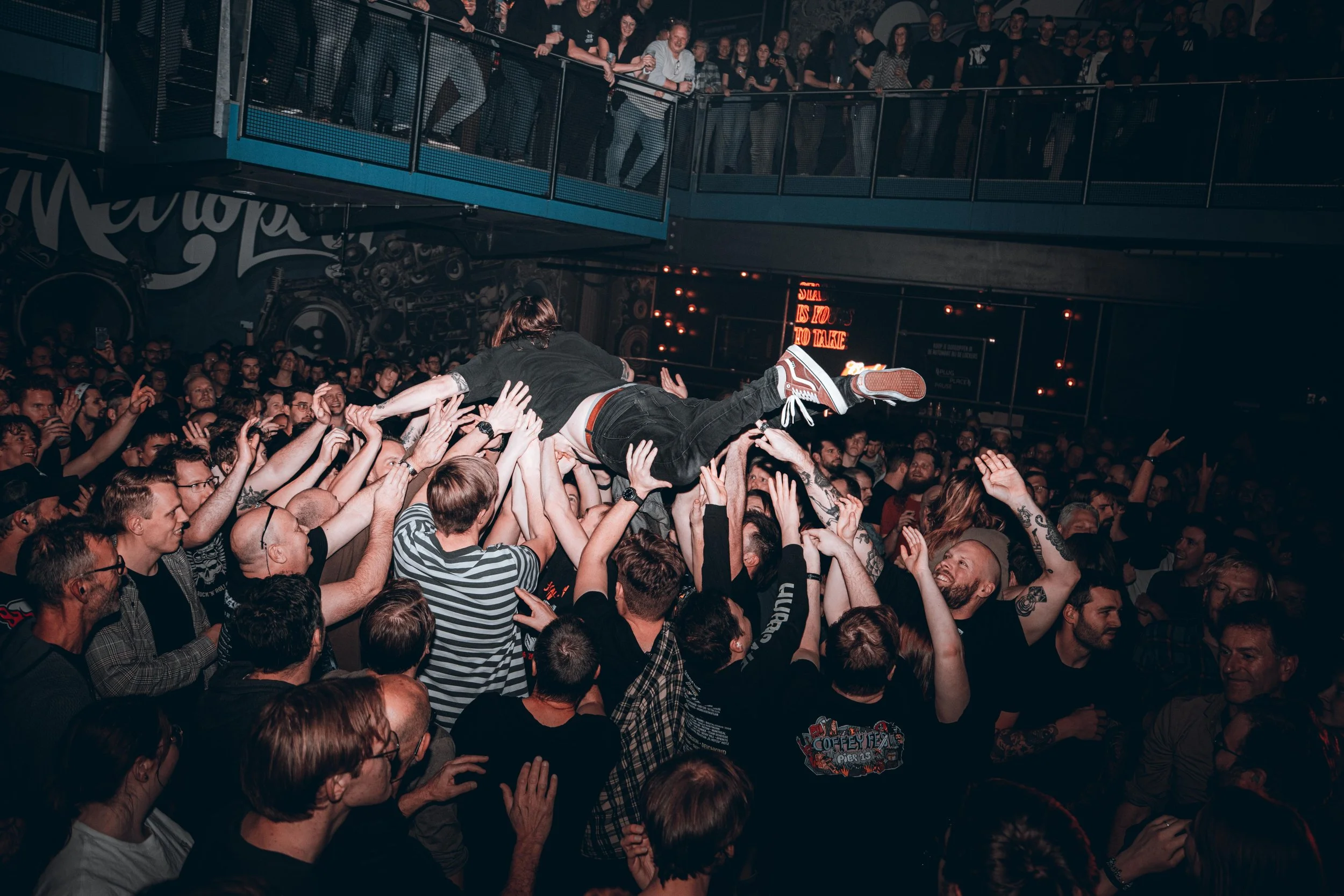 A person crowd surfing at a concert, supported by many people, with the audience raising their hands. The venue is crowded with a second level balcony filled with onlookers.