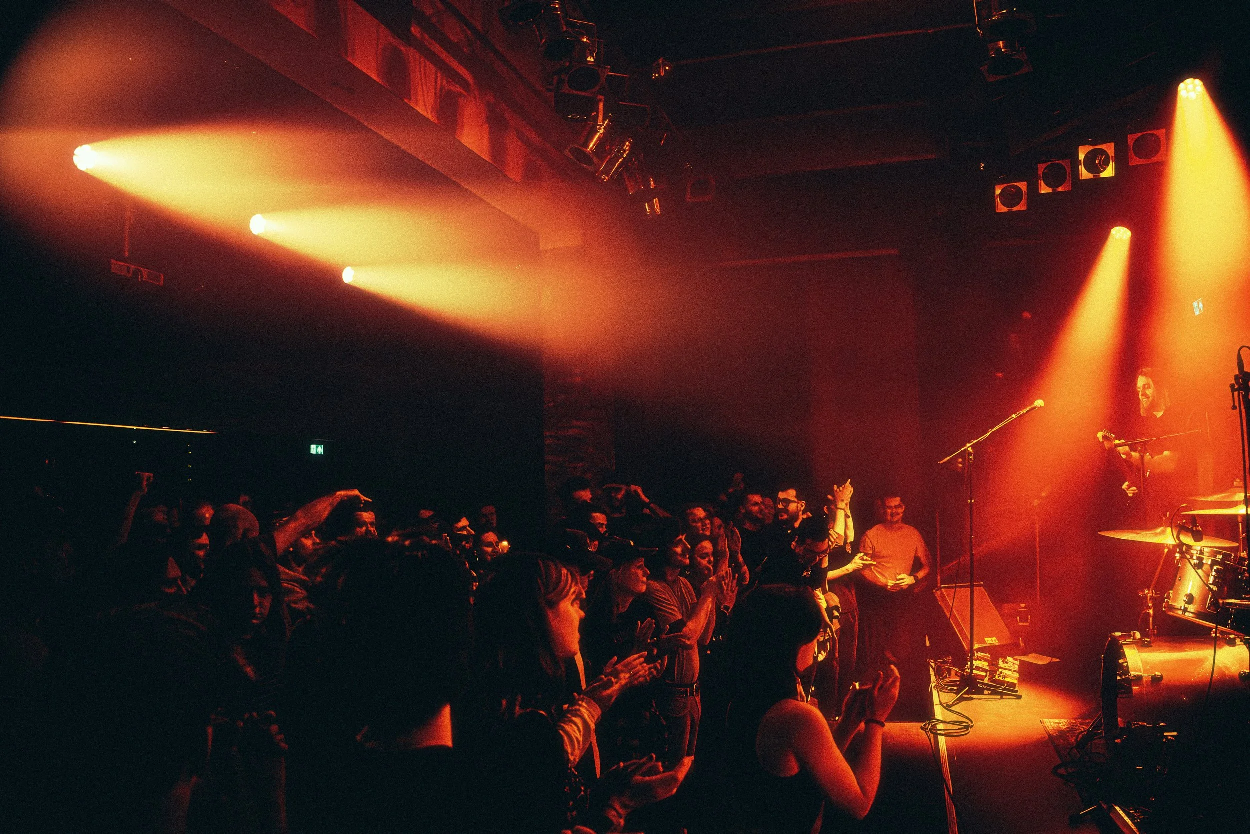 Audience enjoying live music performance in a dimly lit venue with orange stage lighting and a musician on stage playing guitar.