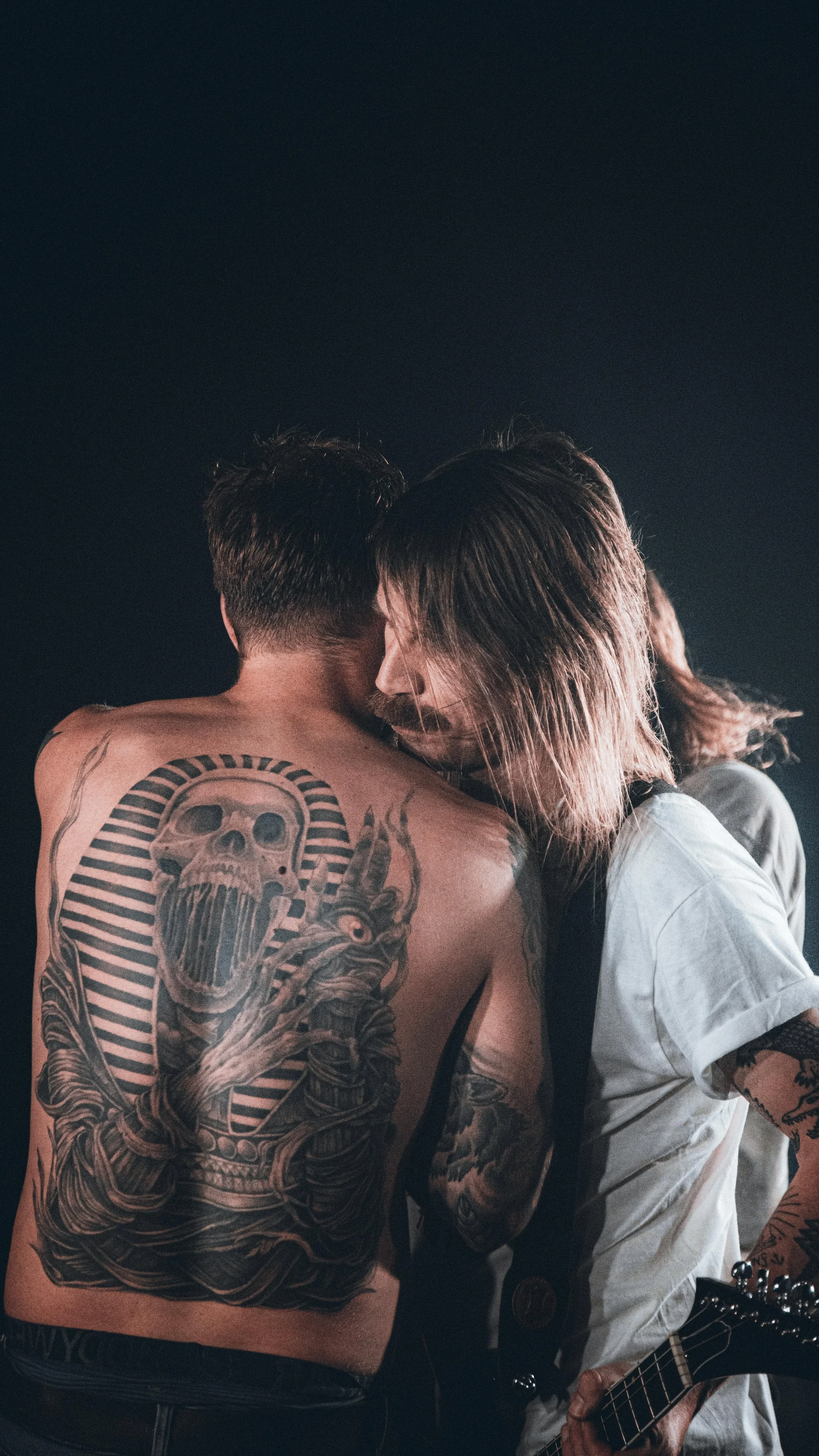 Two men embrace passionately, one with tattoos on his back including a prominent skull and striped pattern, the other with long hair and holding a guitar.