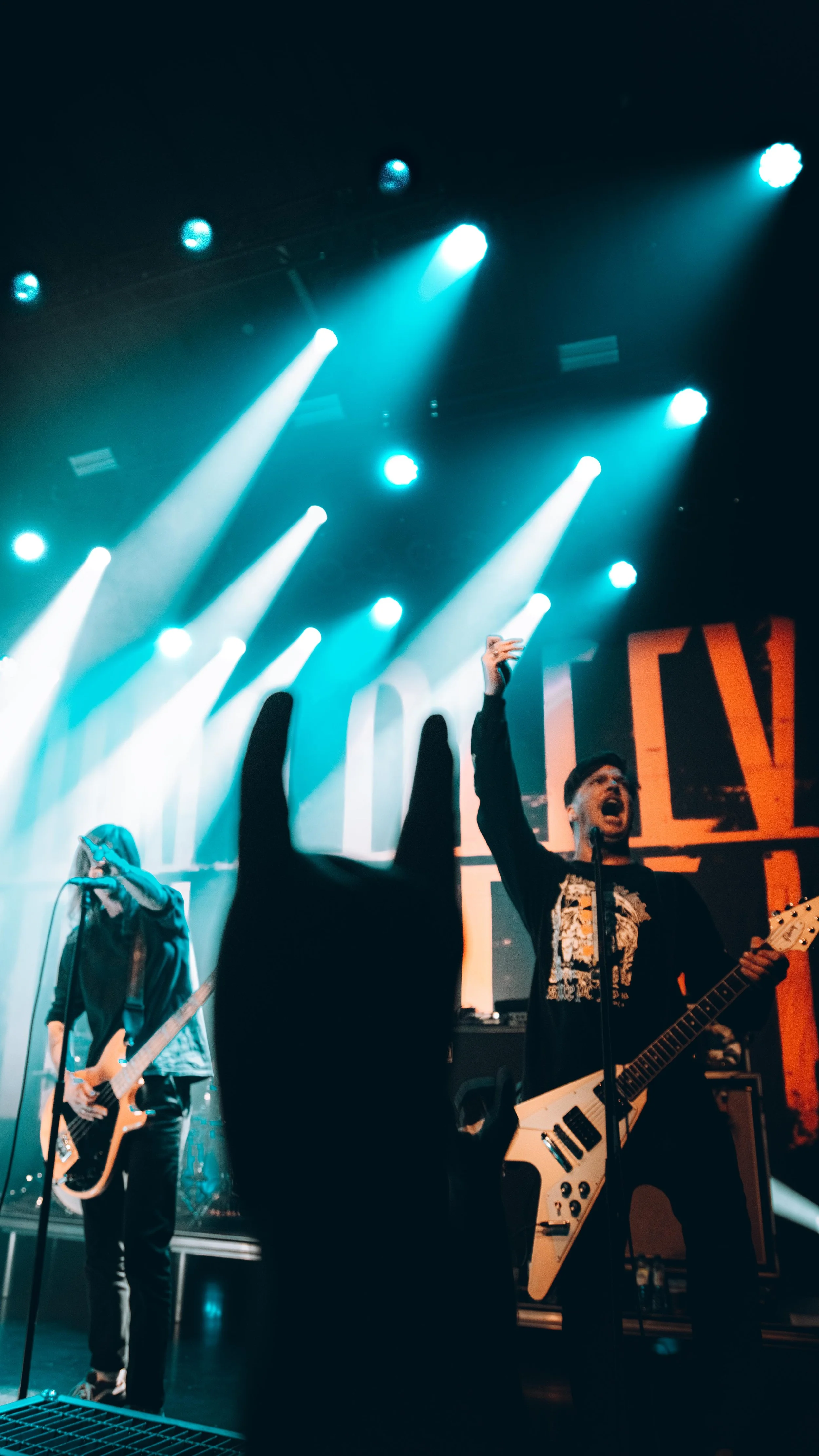 Musicians performing on stage during a concert, with bright stage lights overhead, including a guitarist singing into a microphone and a bassist behind, audience hand visible in the foreground.