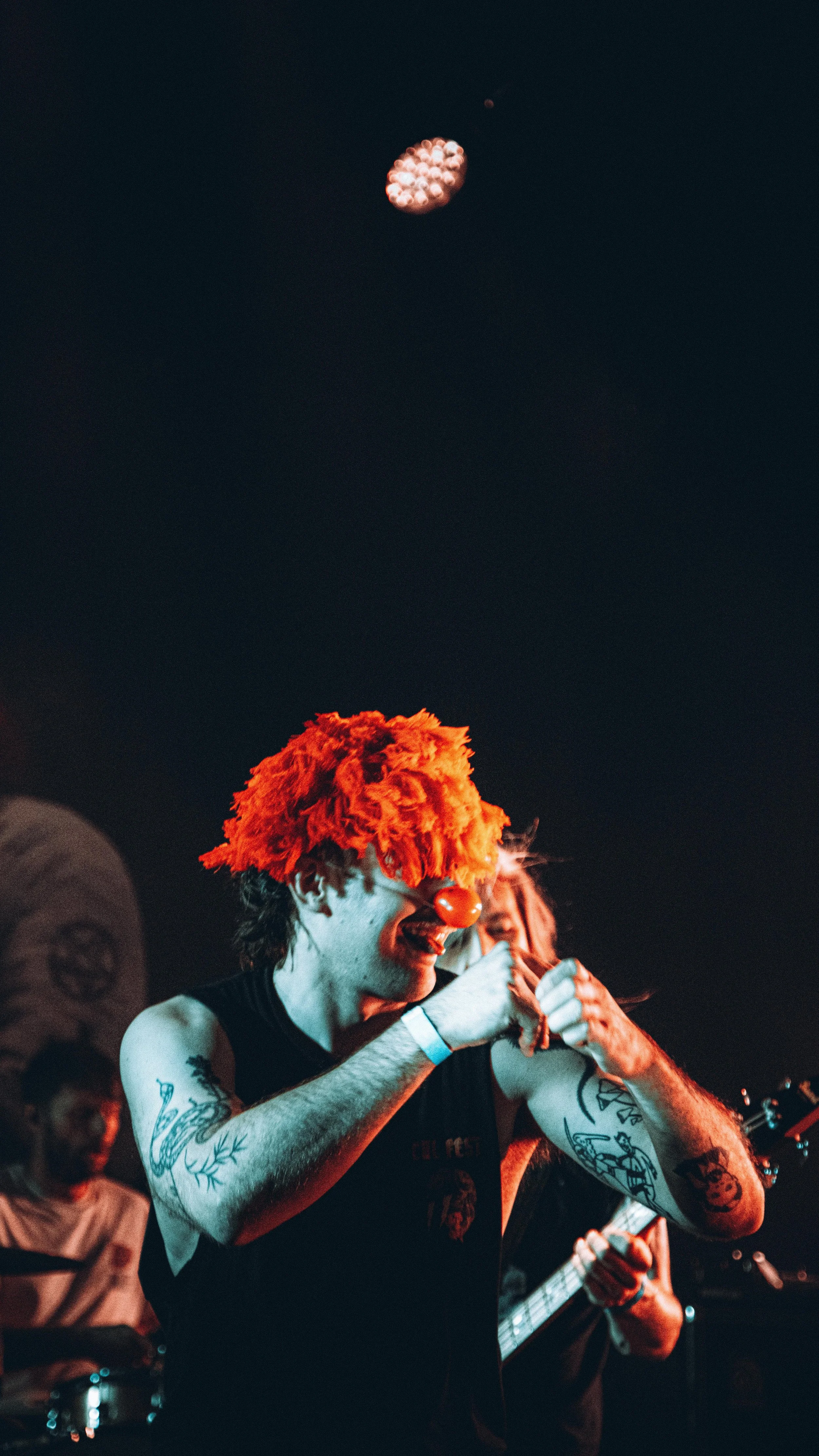 Person with orange wig and clown nose celebrating during a concert, holding a microphone, with a guitarist in the background.