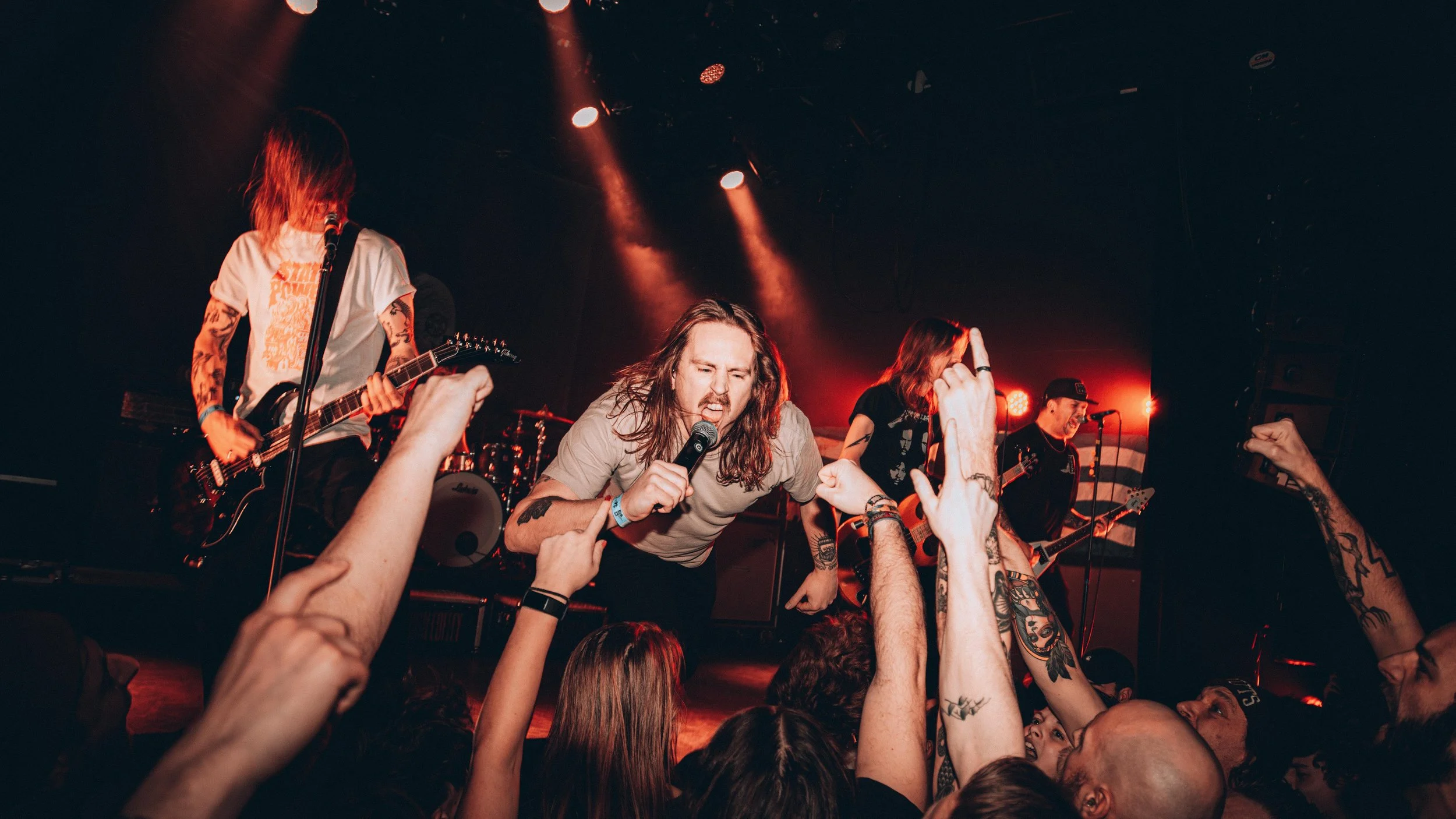 A band performs live on stage with a lead singer leaning into the crowd, surrounded by fans with raised hands, in a dimly lit concert venue.