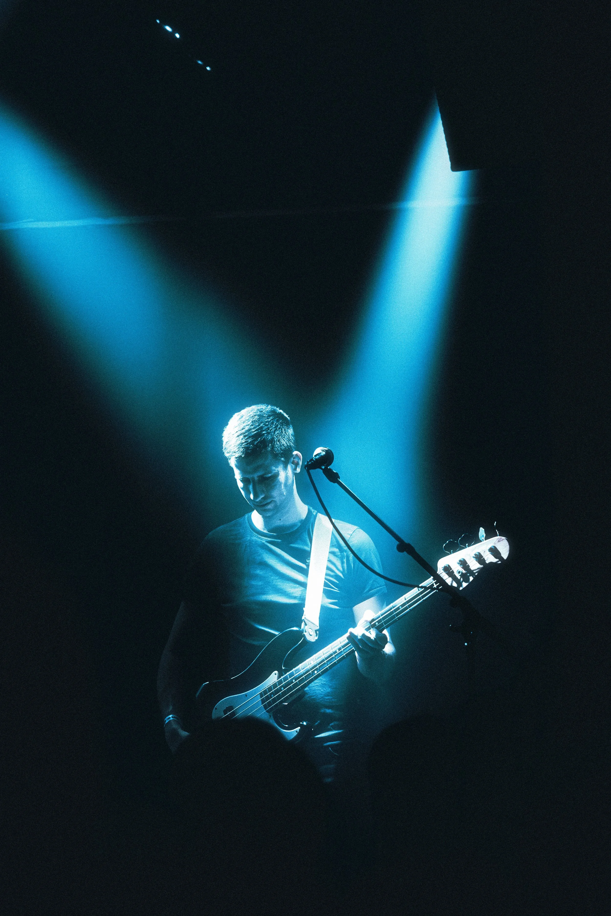 A young man playing an electric bass guitar on stage, illuminated by blue stage lights, with a microphone in front of him.