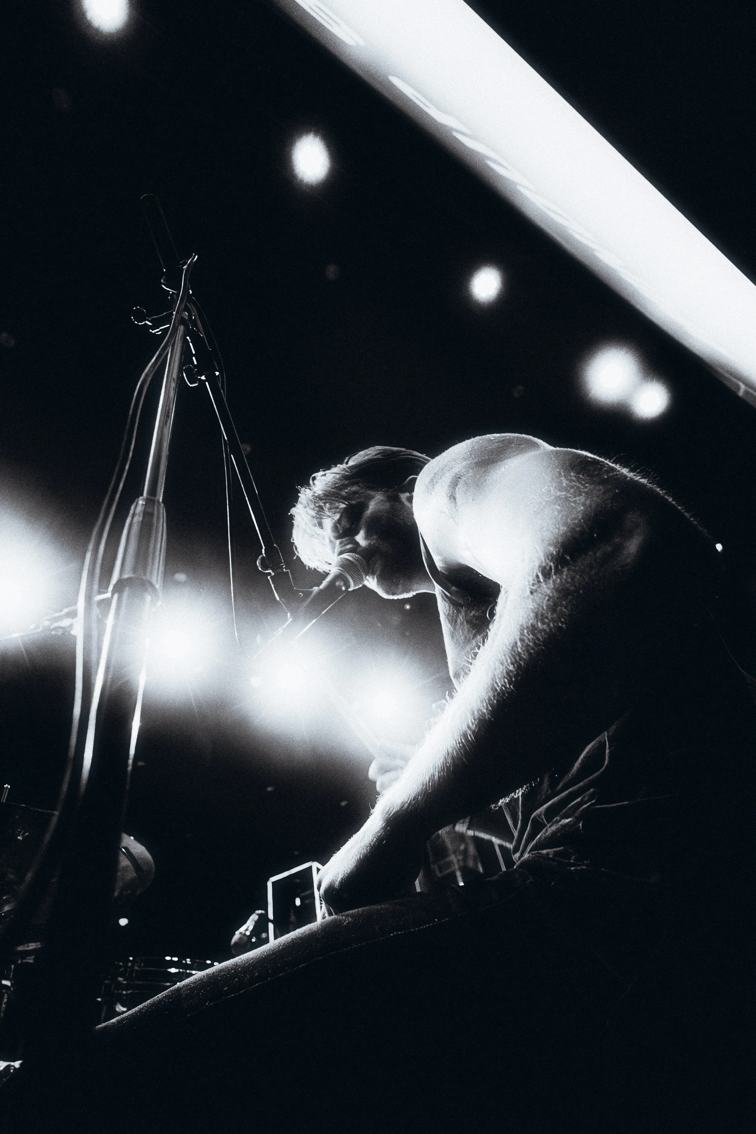 Black and white photo of a musician singing into a microphone on stage, with stage lights overhead.
