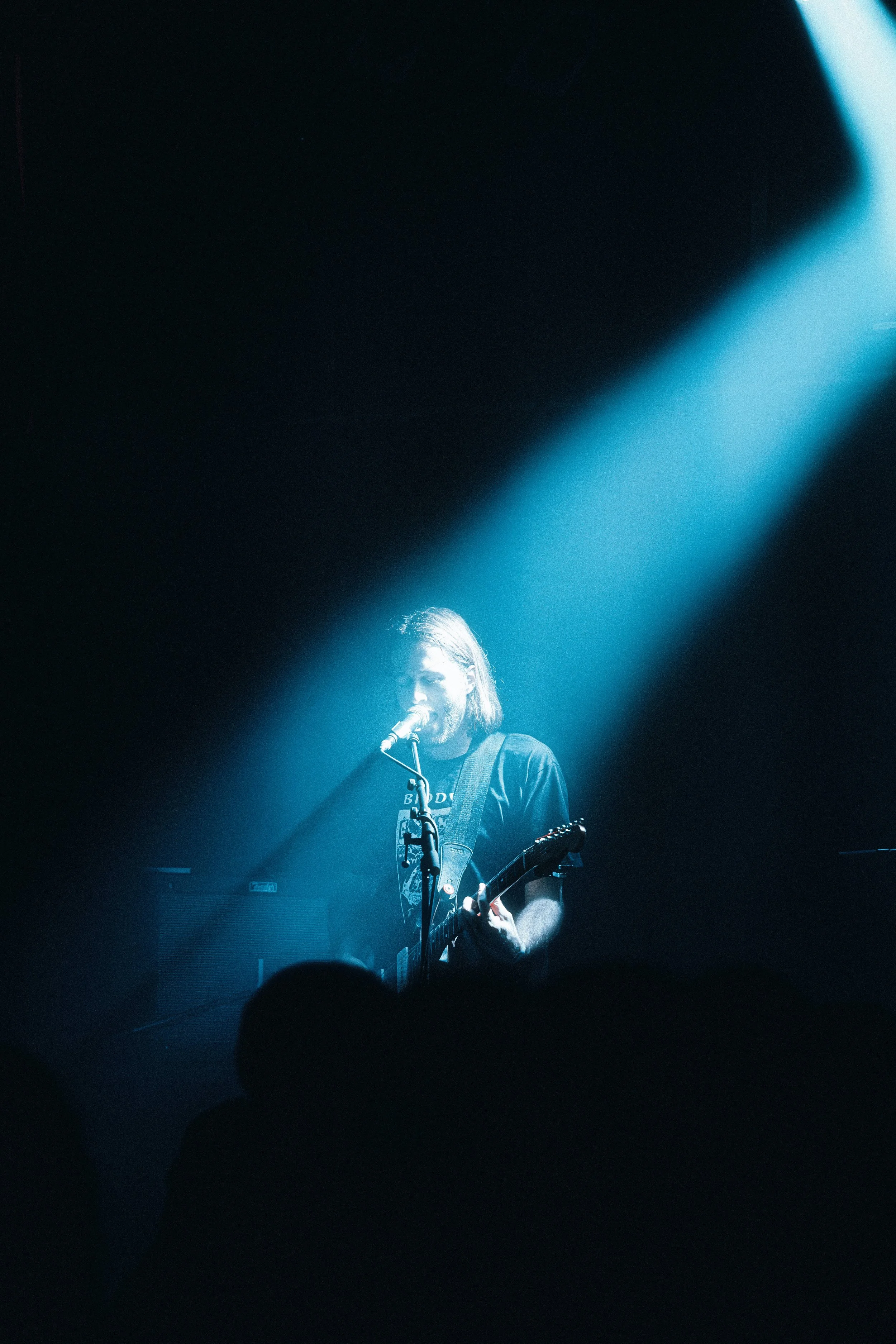 A musician performing on stage, playing an electric guitar and singing into a microphone, illuminated by a blue spotlight.