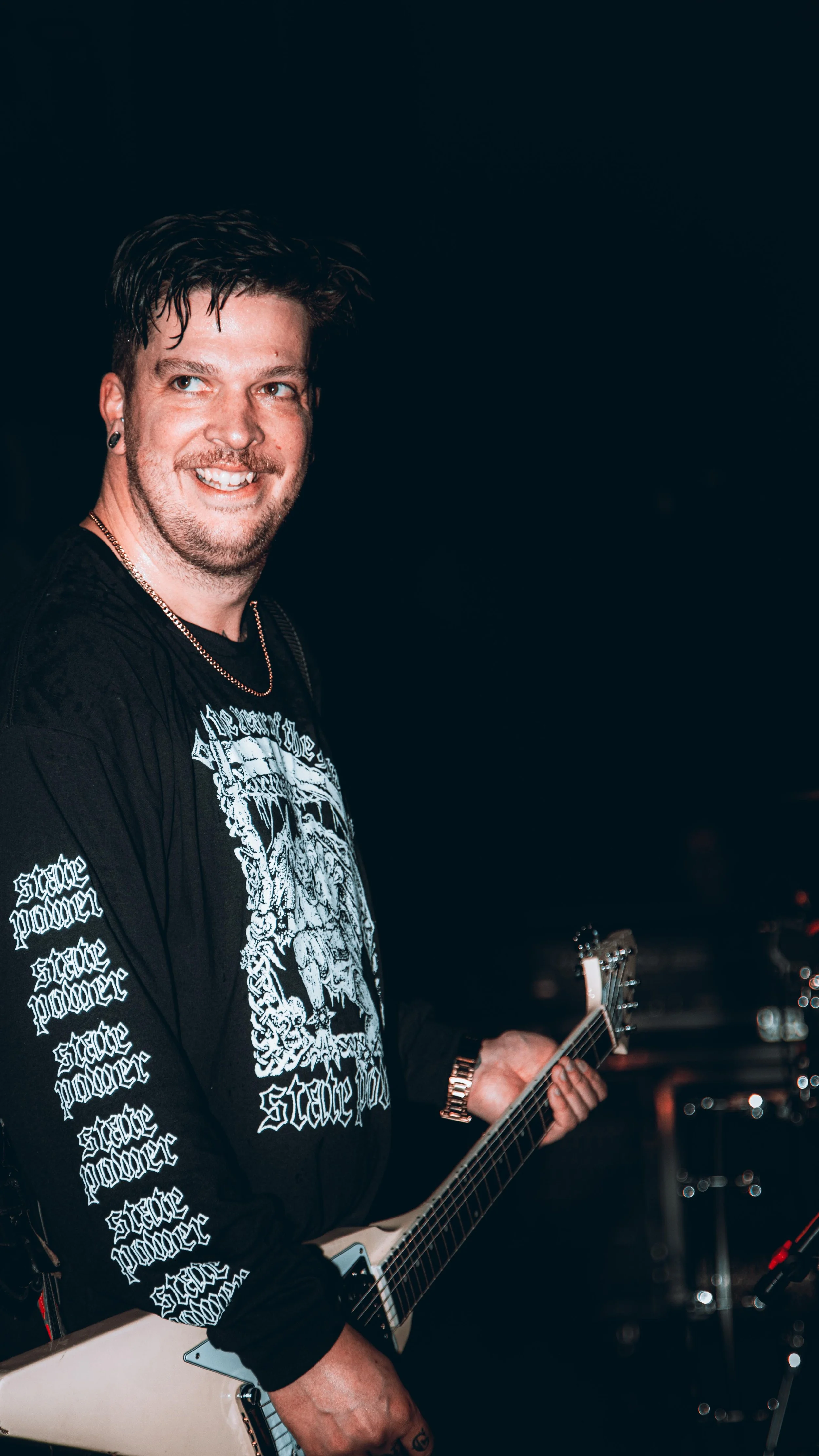 A smiling man playing an electric guitar on stage, wearing a black long-sleeve shirt with white text and design, dark hair styled upward, and a watch on his left wrist.
