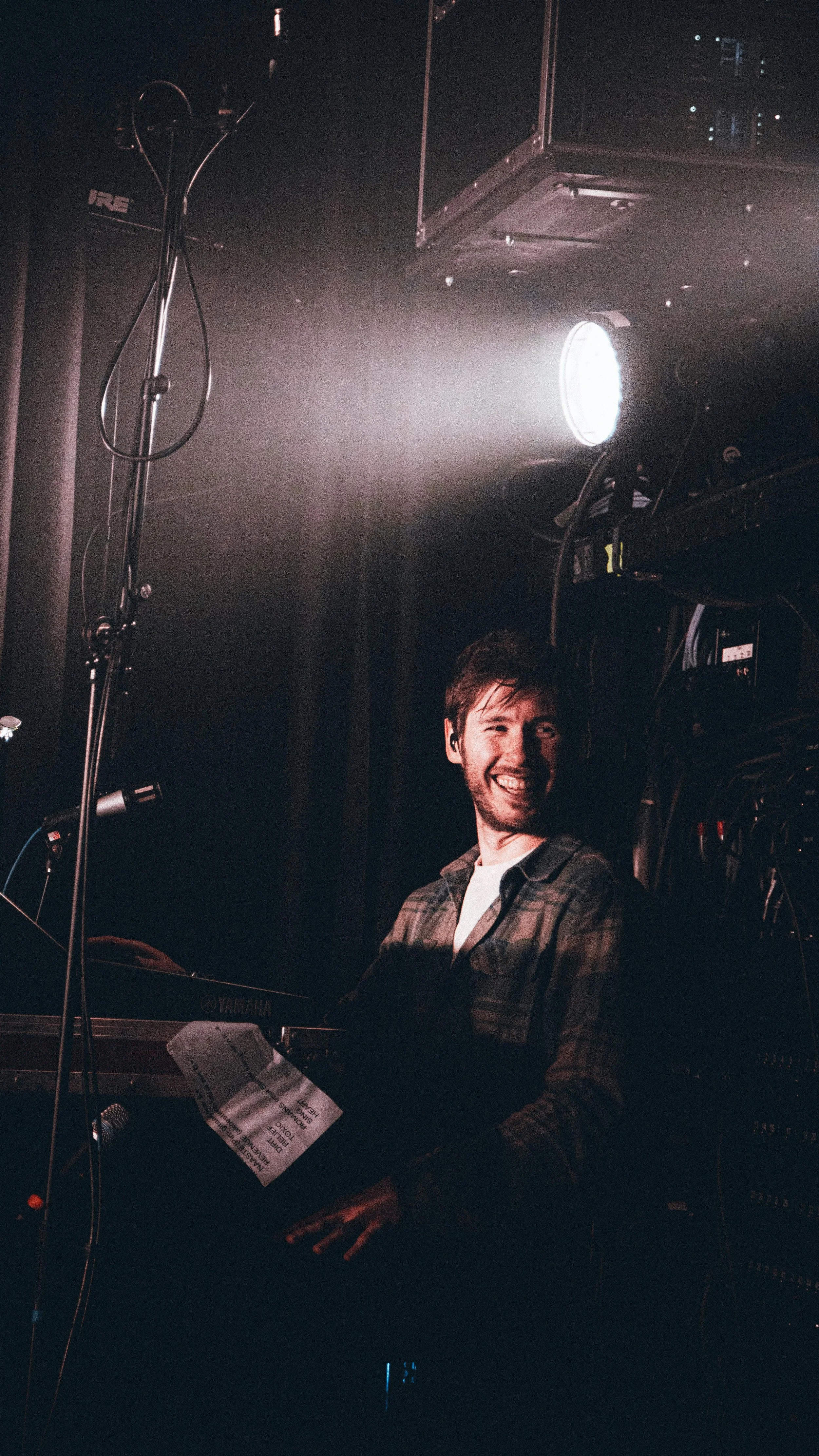 A man smiling and playing keyboard on stage with lighting equipment around him.