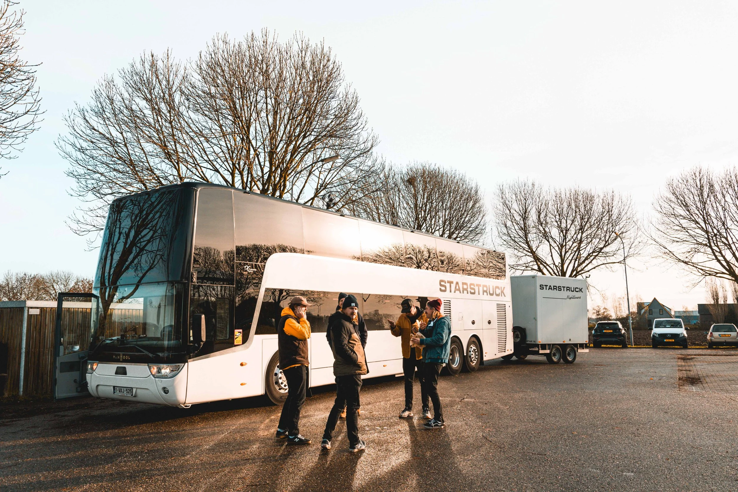 Group of five people standing near a white double-decker bus in a parking lot, engaged in conversation, with leafless trees and parked cars in the background.