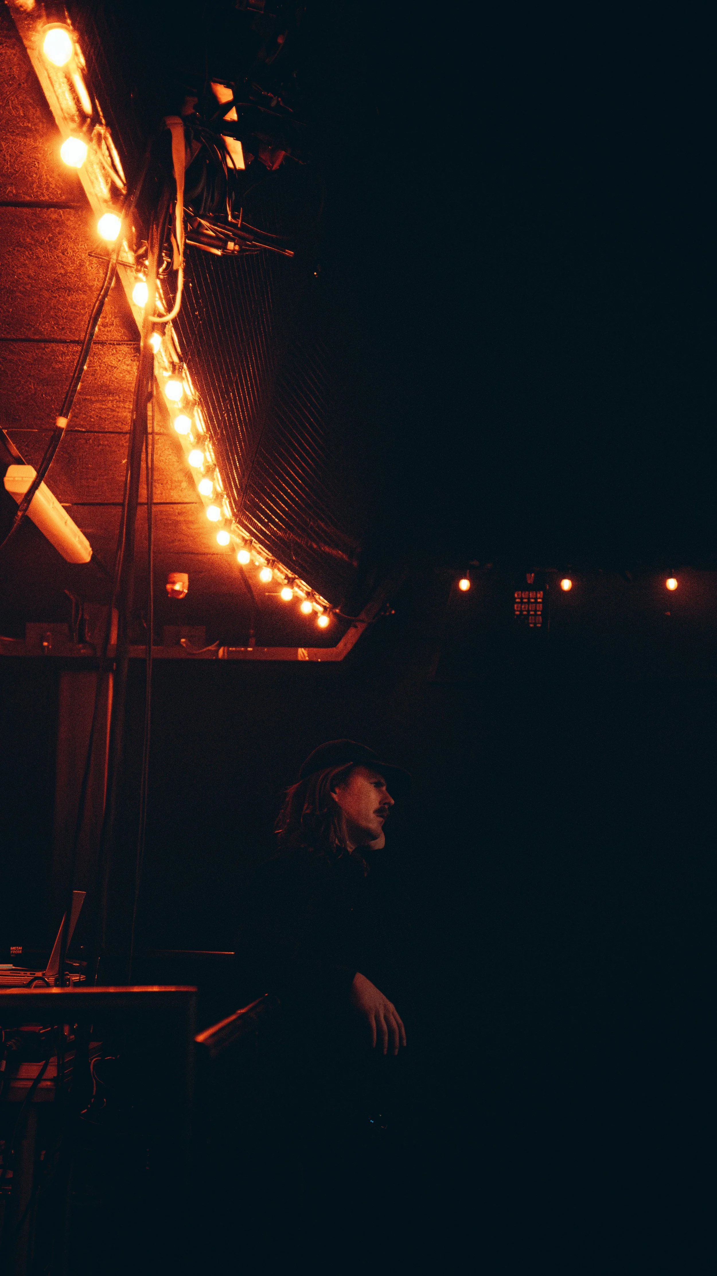 A person with long hair and a hat sitting next to a soundboard or DJ equipment in a dark, dimly lit space with orange string lights hanging above.