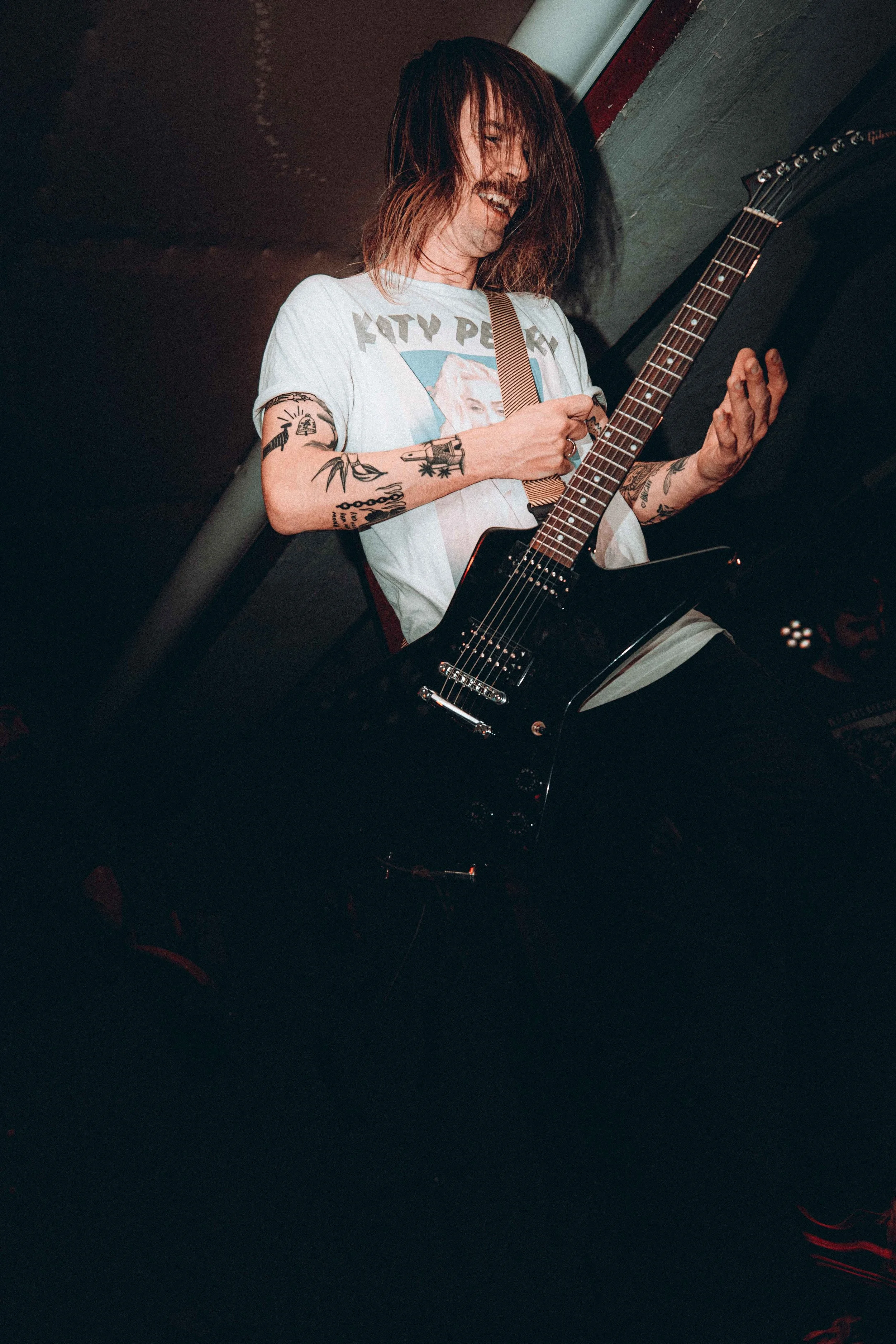Man with long hair and tattoos playing an electric guitar on stage, wearing a mostly white T-shirt.